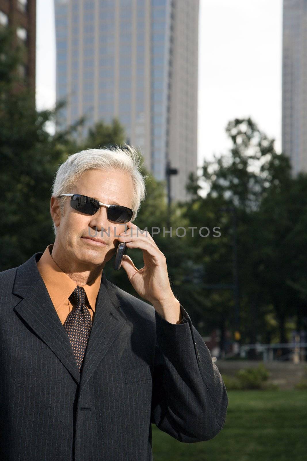 Caucasian middle aged businessman in sunglasses holding cell phone outdoors.