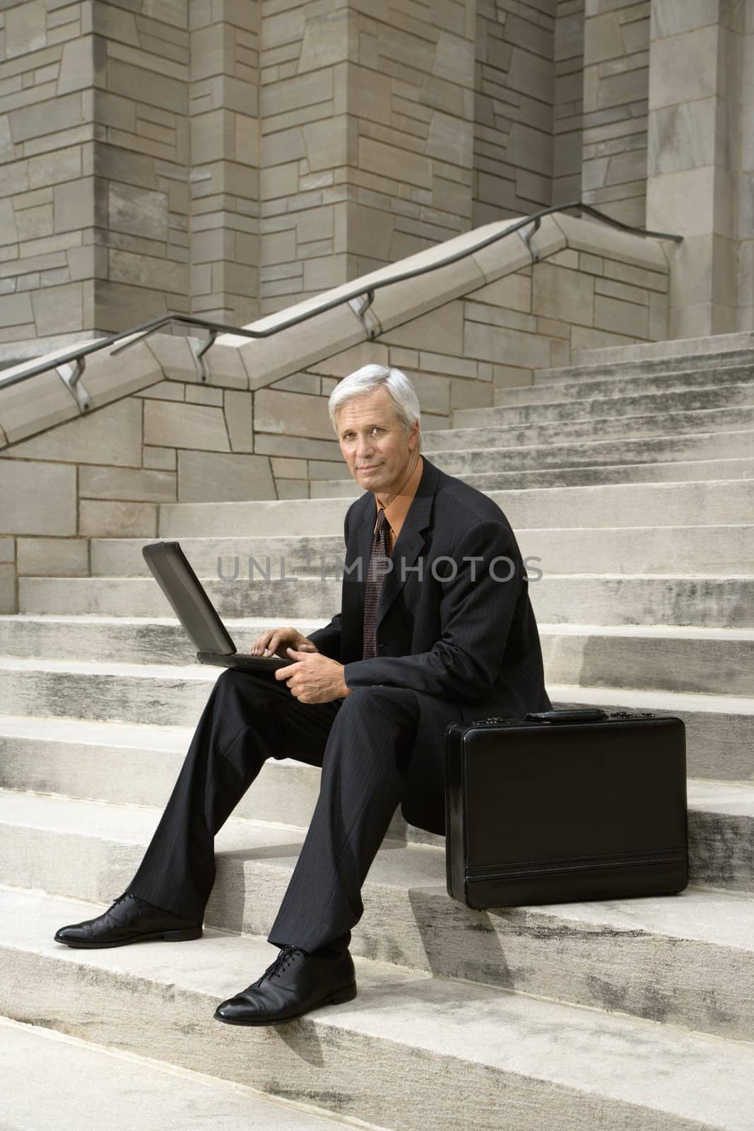 Caucasian middle aged businessman sitting on steps outdoors with laptop and briefcase.