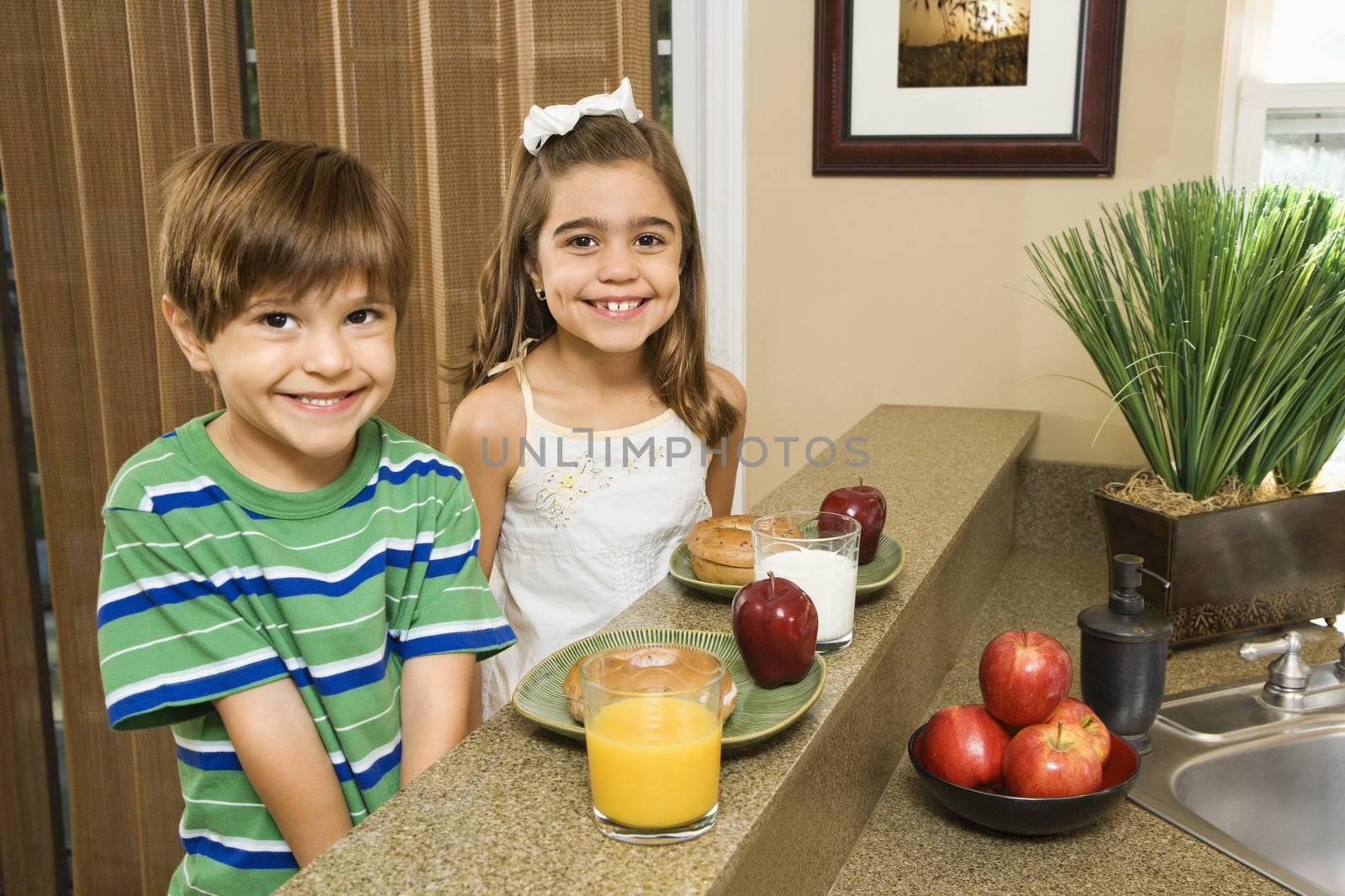 Young Hispanic brother and sister sitting at kitchen bar with healthy breakfast smiling at viewer.