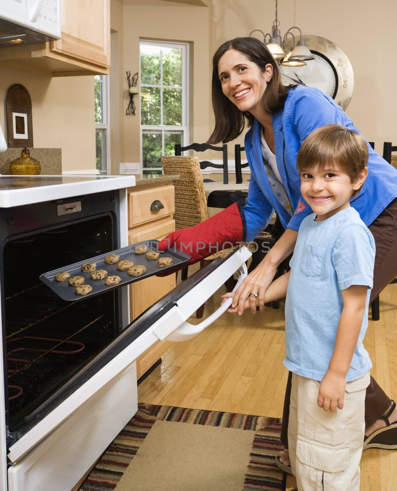 Hispanic mother and son putting cookies into oven and smiling at viewer.