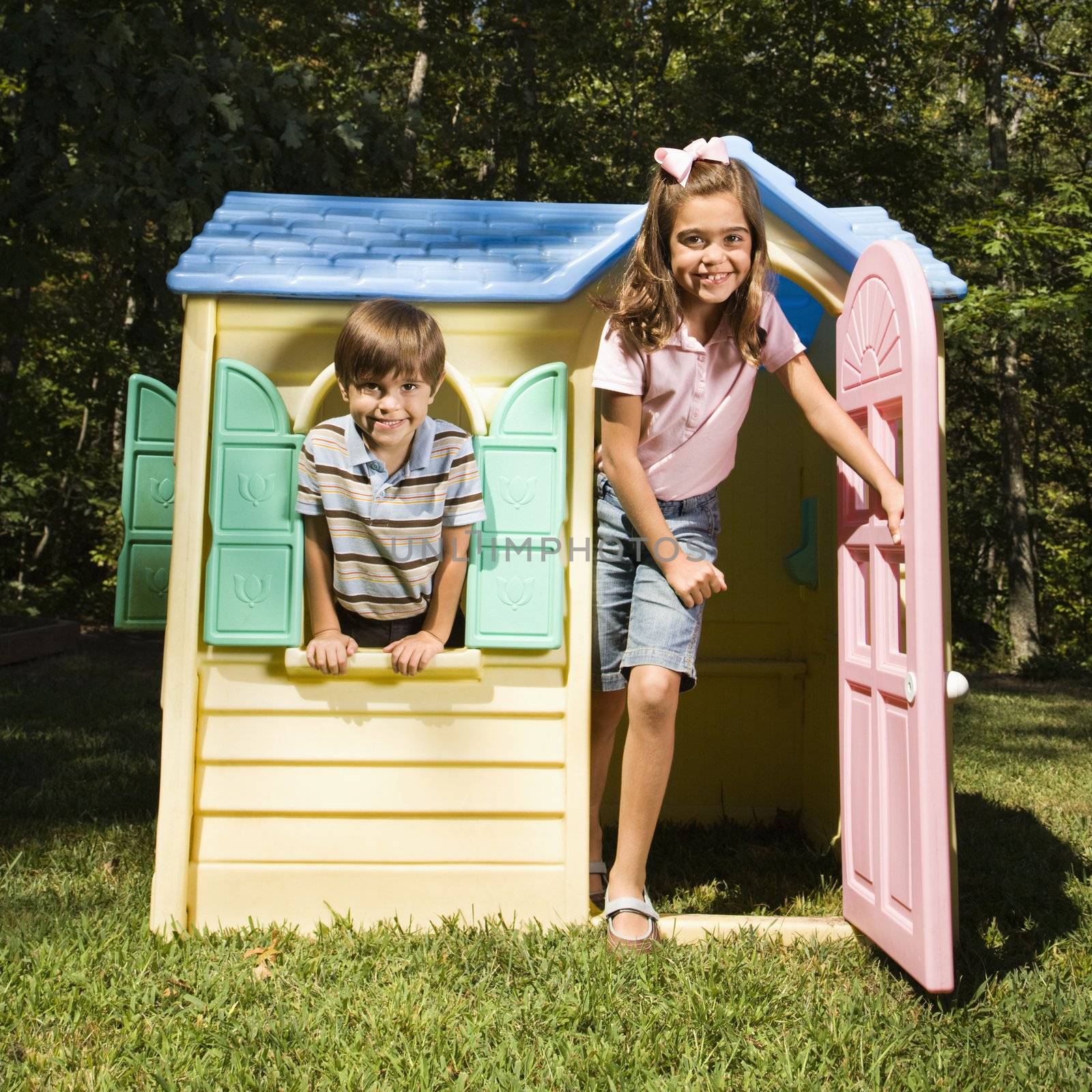 Hispanic boy and girl in outdoor playhouse smiling at viewer.