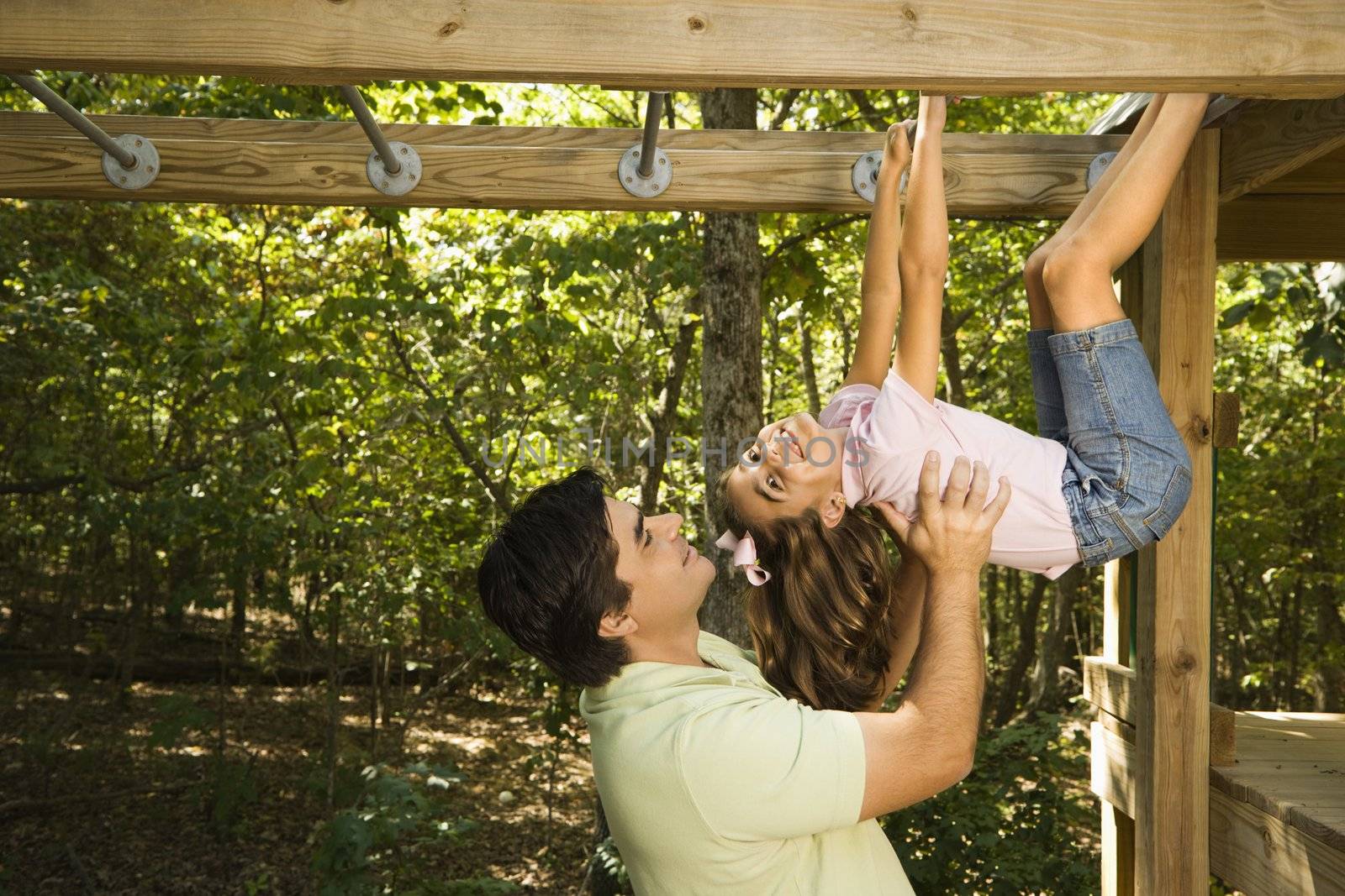 Hispanic girl hanging by arms and legs from monkey bars smiling at father helping her.