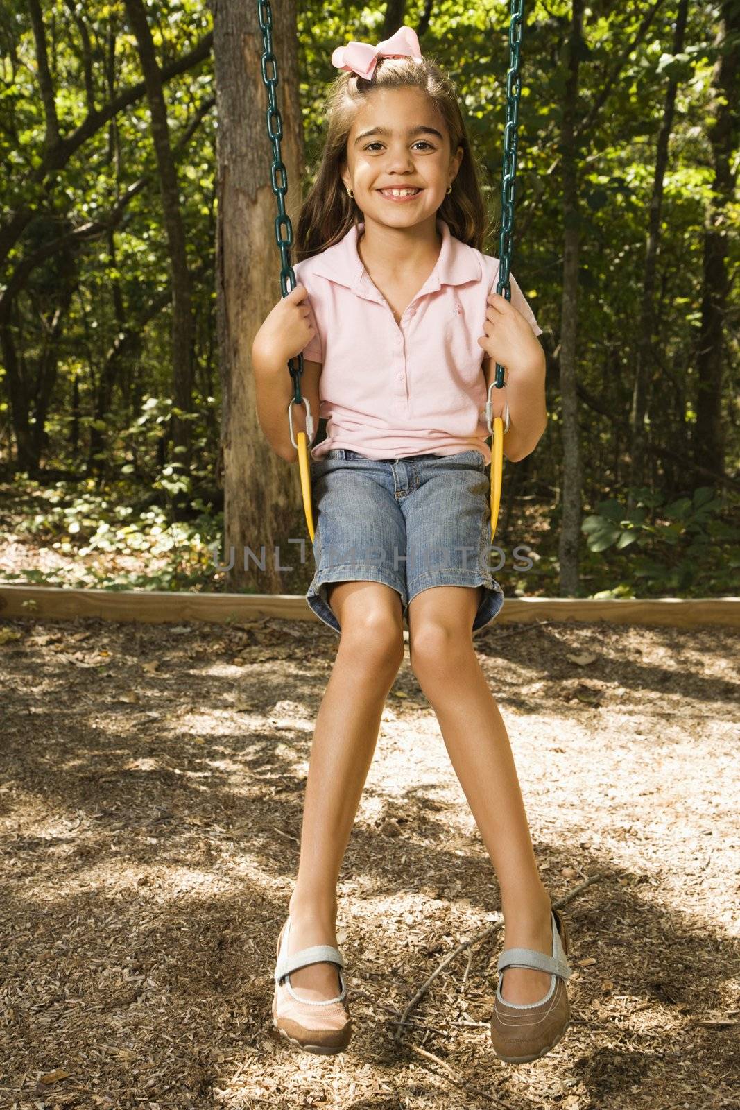 Hispanic girl sitting on playground swing smiling at viewer.