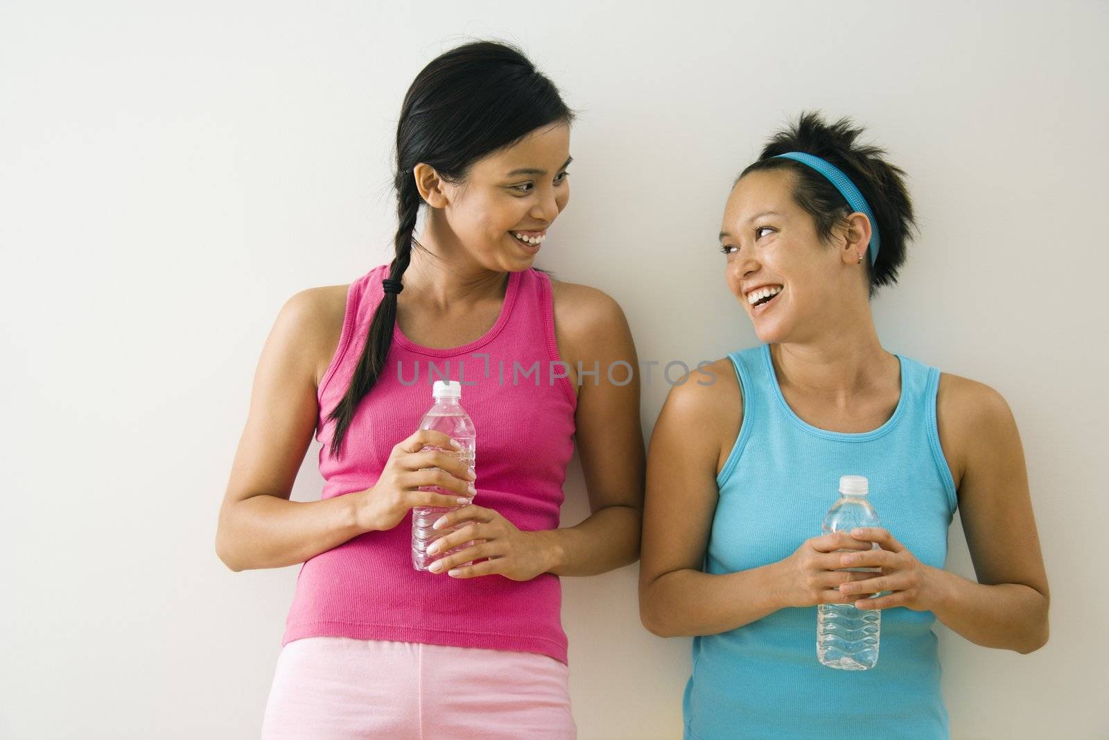 Two young women standing in workout clothes talking and smiling holding bottled water.