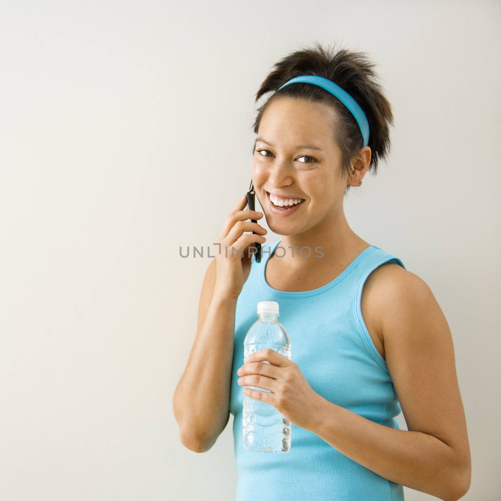 Young woman in fitness clothing holding bottled water talking on cellphone smiling.