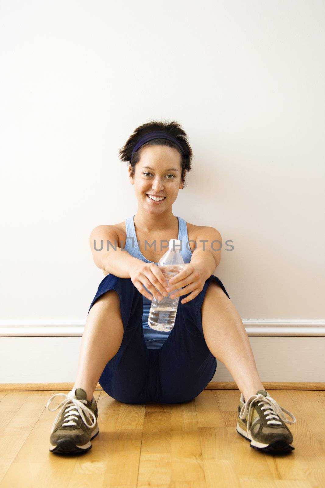 Young woman in fitness outfit sitting on floor with bottled water smiling.