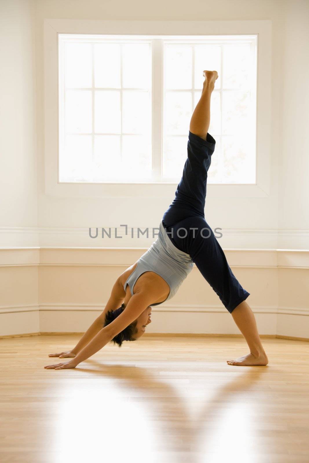 Young woman doing downward dog pose with one leg raised on wooden floor indoors by sunlit window.