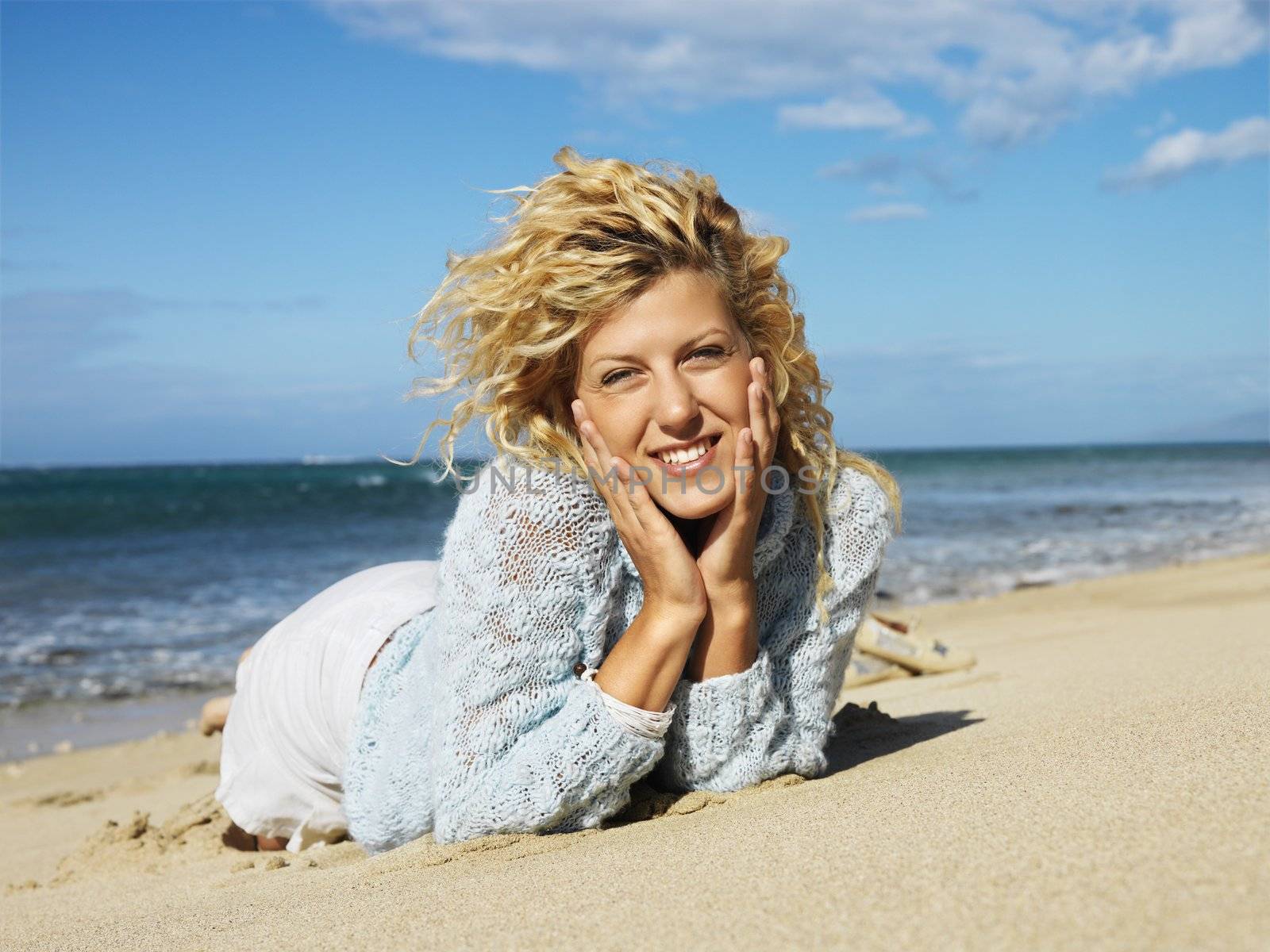 Pretty young blond woman lying in sand on Maui, Hawaii beach with head resting on hands smiling.
