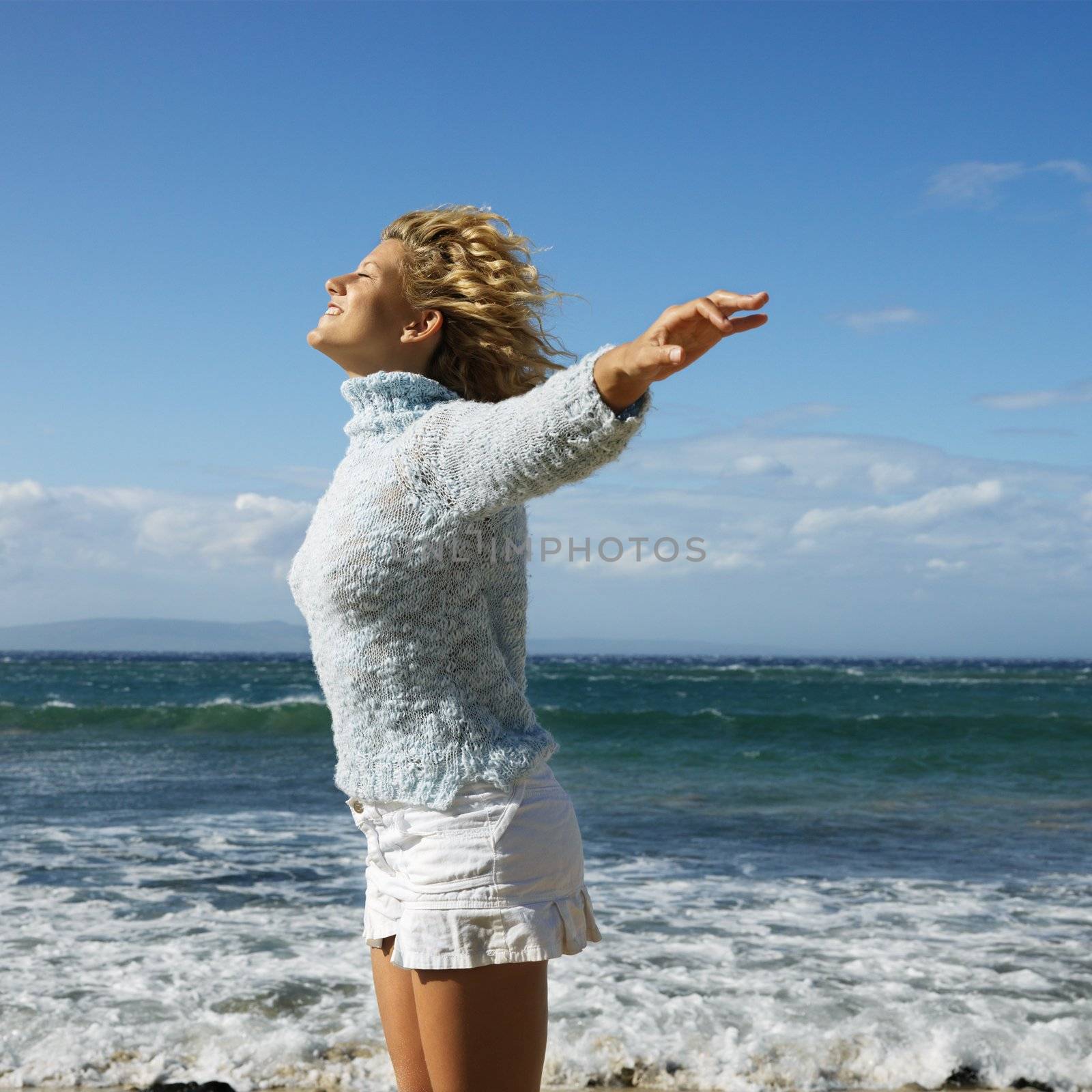 Pretty young blond woman standing on Maui, Hawaii beach with arms out to side and wind blowing hair smiling.