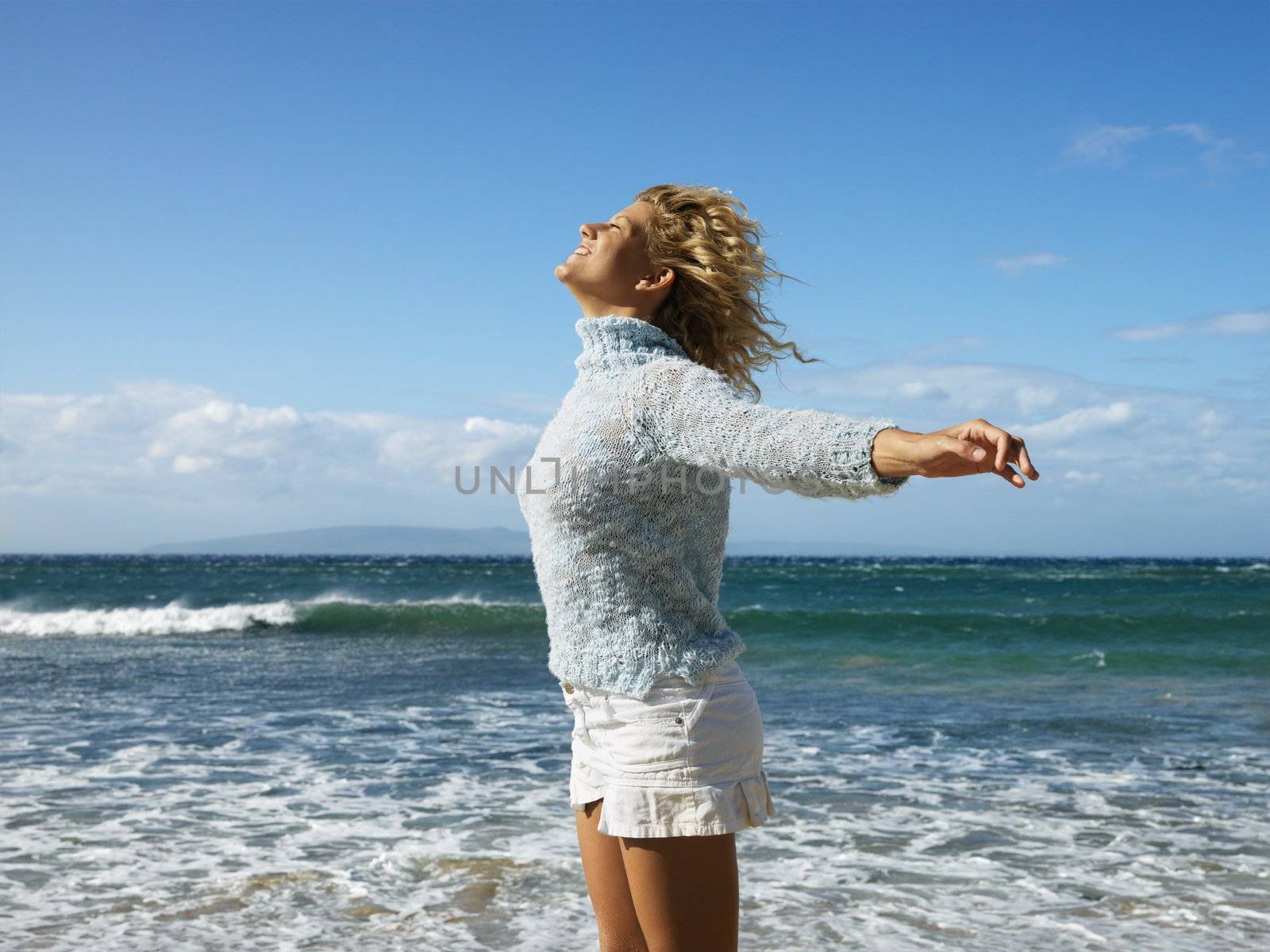 Pretty young blond woman standing on Maui, Hawaii beach with arms out to side and wind blowing hair smiling.