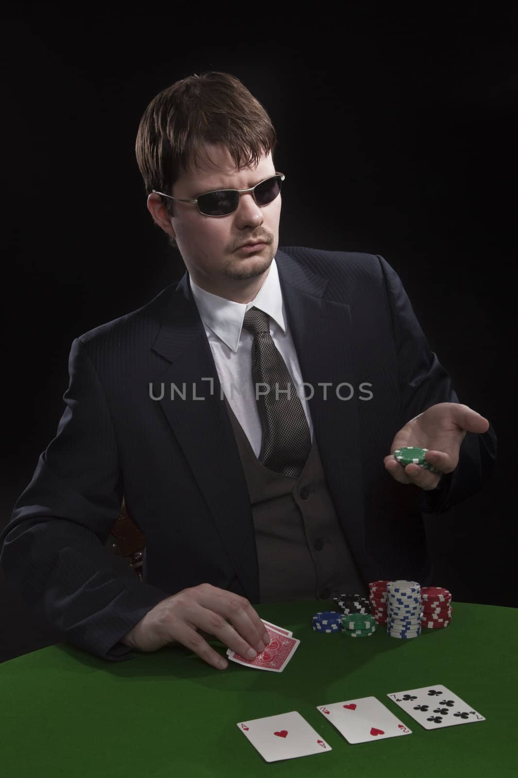 Man in suit with sun glasses playing poker on green table. Chips and cards on the table.