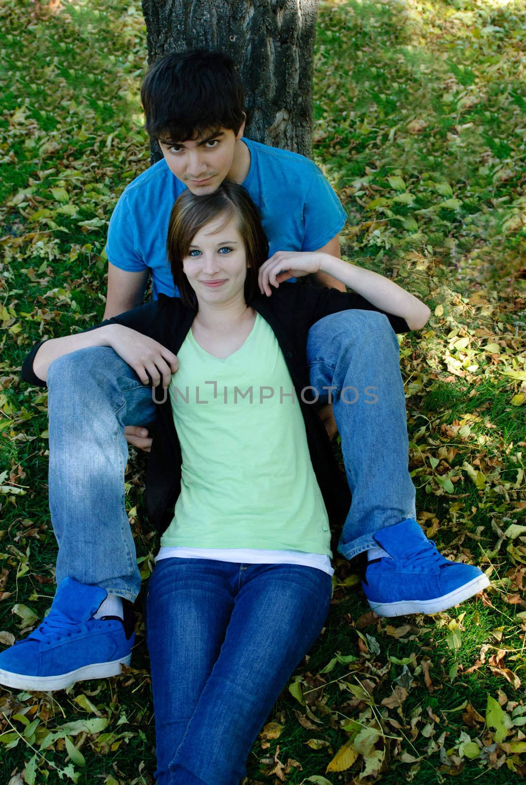 A teenage couple is resting by a tree in the fall with leaves on the ground