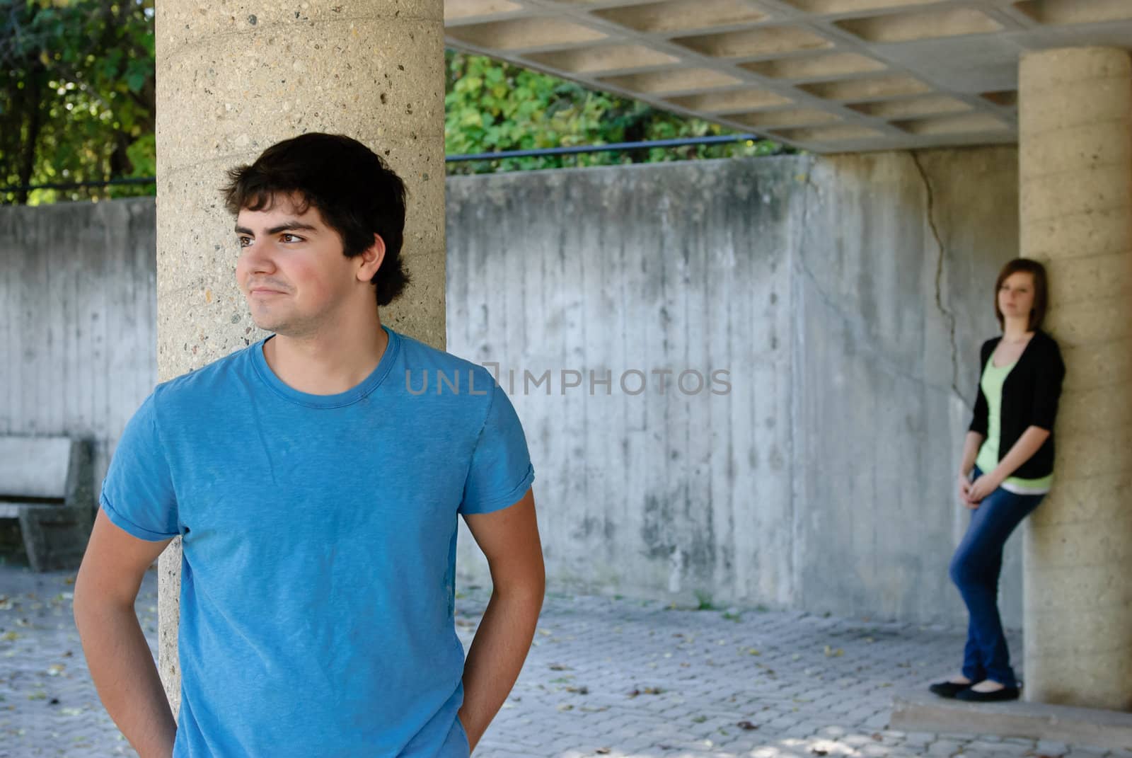 A male teenager is posing by a stone pillar with his girlfriend in the background