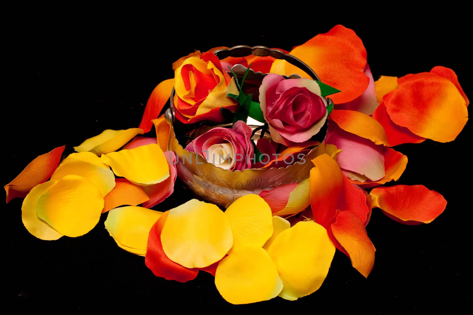 A silver basket with three artificial roses on orange, yellow and pink rose textile petals on black
