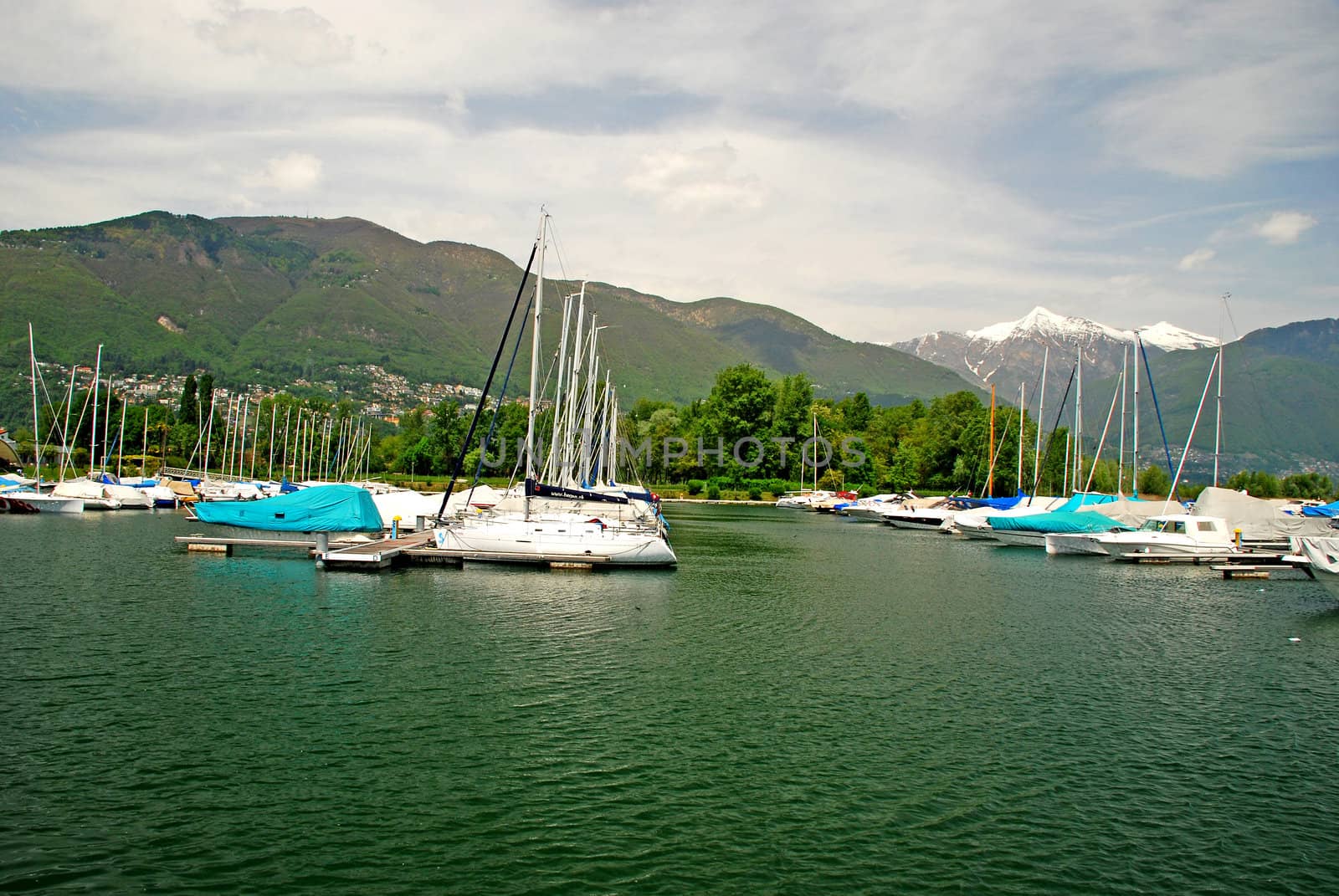 There is a sailing port and yacht club in Ascona, in Lake Maggiore. Behind yachts, there is a park,  at the background ther is a part of Locarno city and Alps with snow covered peaks of   Trosa and Madone mountains and cloudy sky