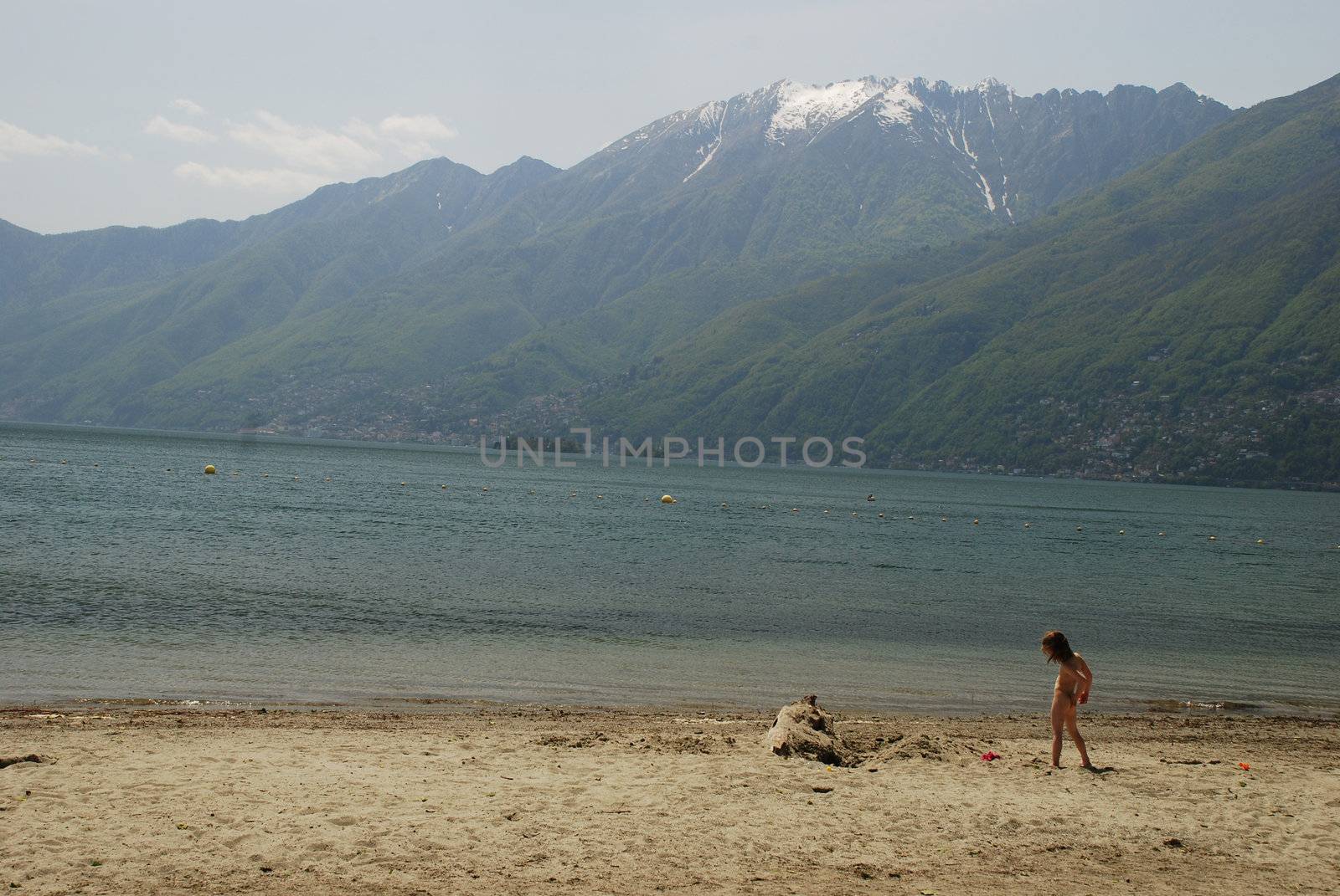 There is a Ascona beach in Lake Maggiore in the middle of May, there is a child playing in the beach, there is a beutiful view in the lake, porto Ronco and  Brissago situated in the border of the Lake, beutiful Leone hill still covered with snow and the cloudy sky