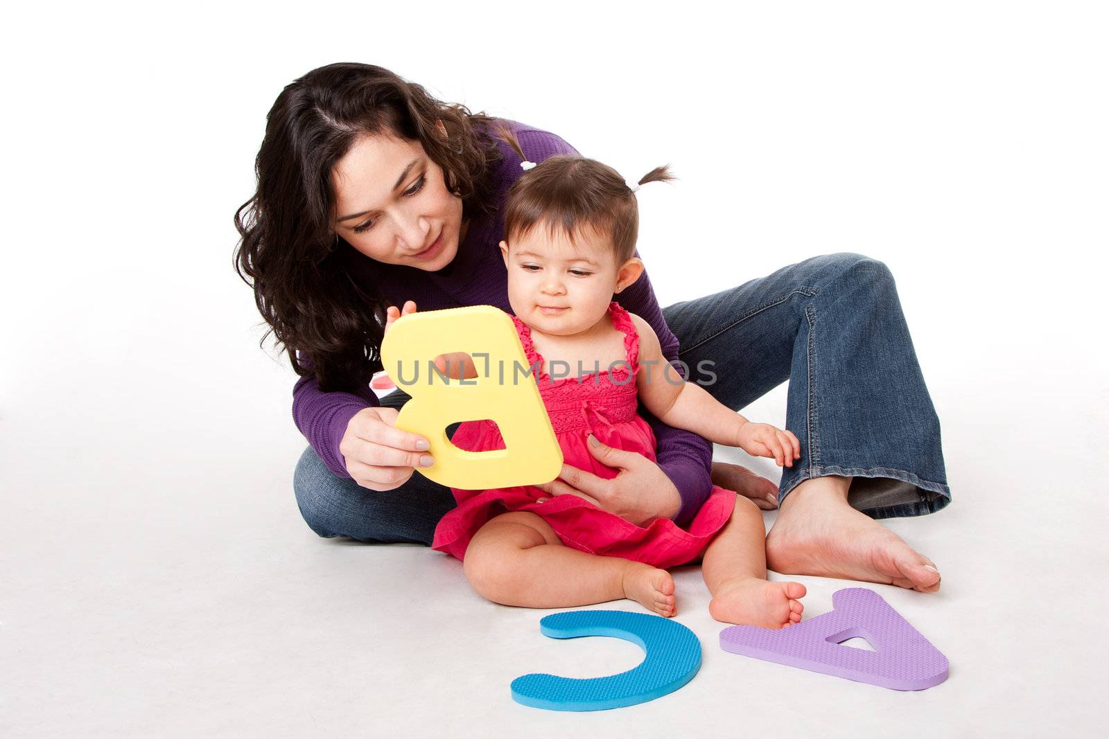 Mother, nanny, or teacher teaching happy baby to learn alphabet, A, B, C, with letters in a playful way, while sitting on floor.