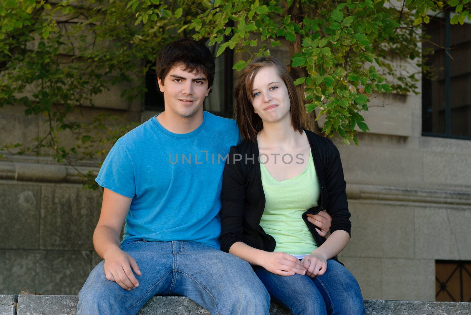A happy teen couple sitting beside each other with a stone building in the background.