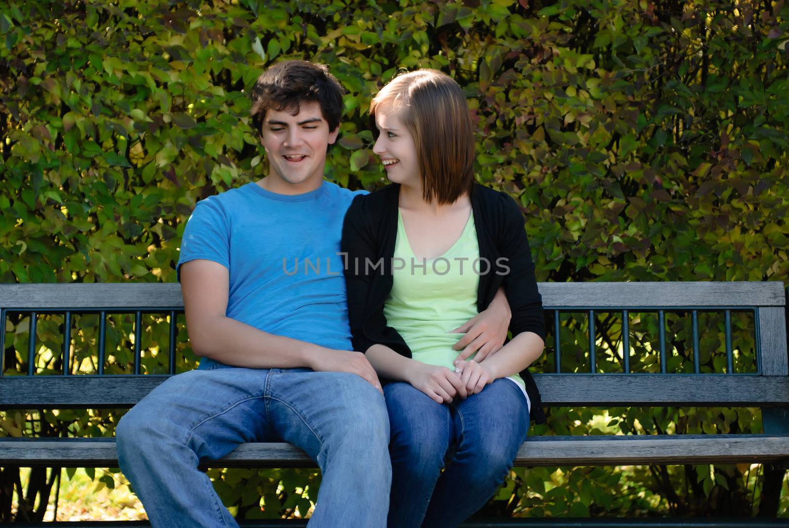 Two teenagers snuggling and sitting on a park bench with leaves in the background.