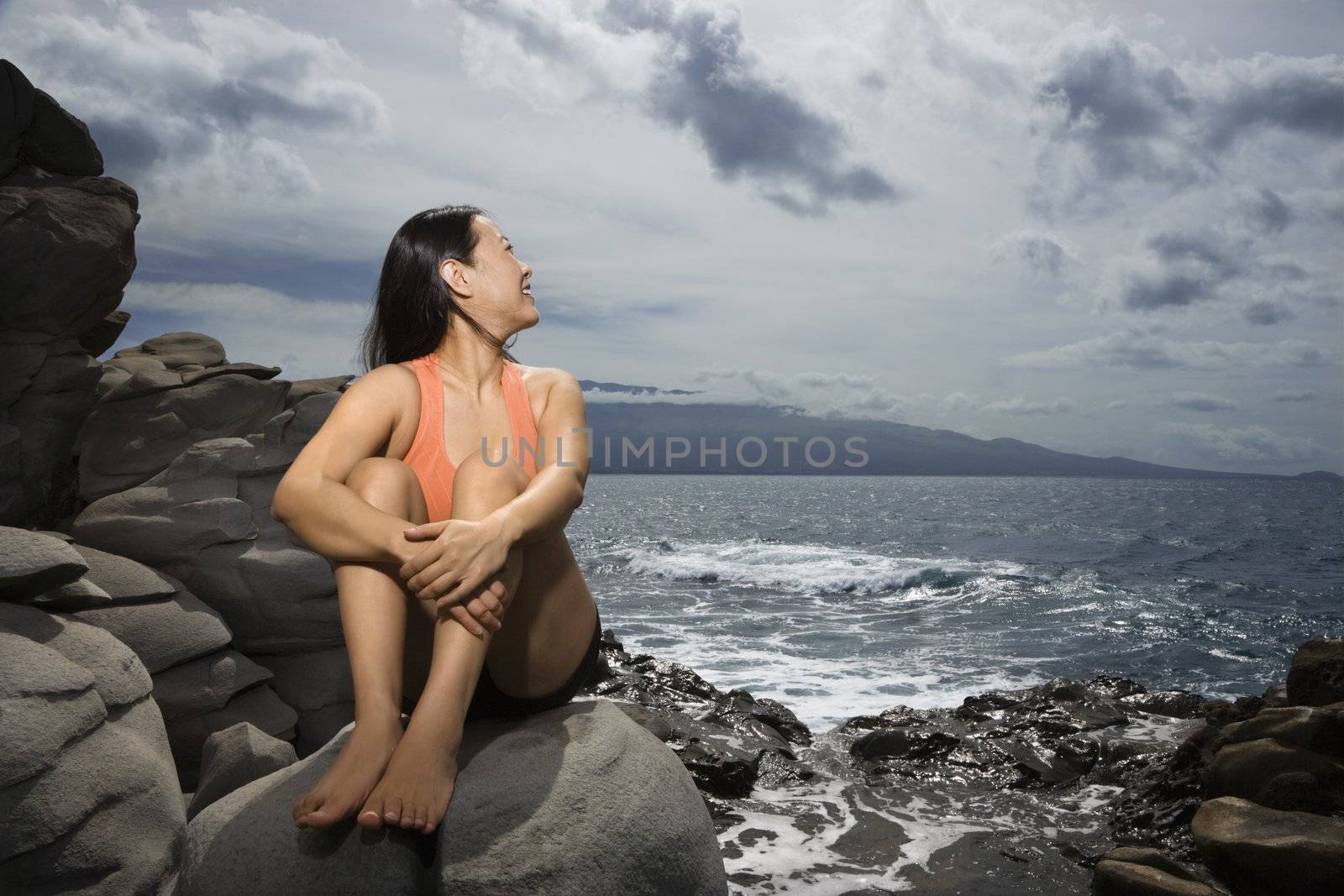 Asian woman sitting on rock by ocean looking over shoulder smiling in Maui, Hawaii