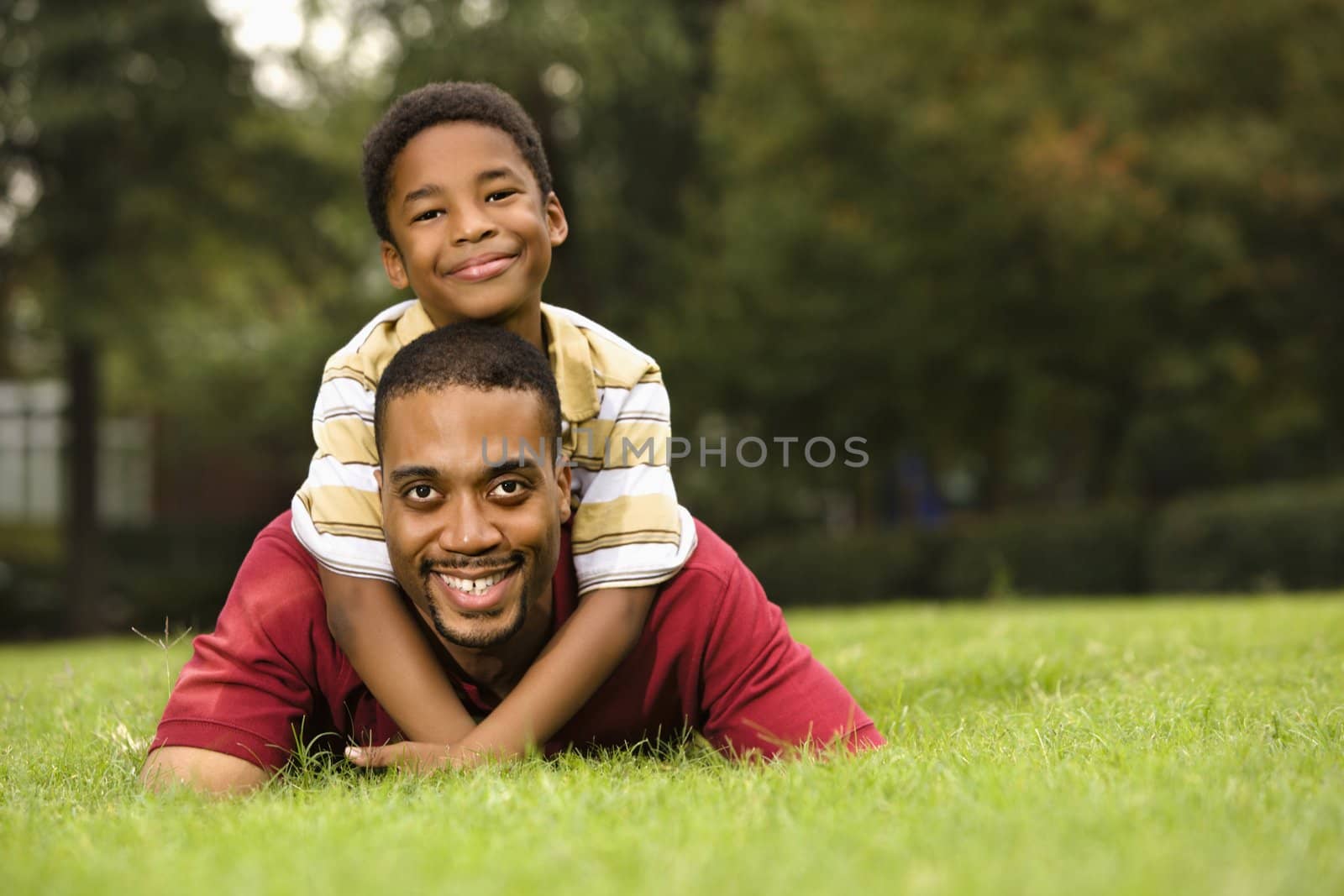 Father lying in grass smiling as son climbs on his back and hugs his neck.