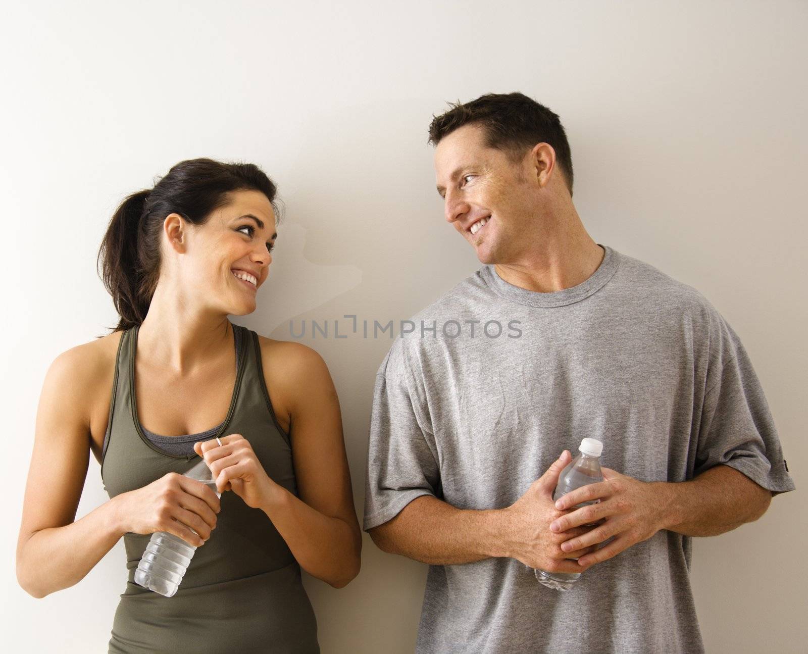 Man and woman at gym in fitness attire holding water bottles standing against wall smiling at eachother.