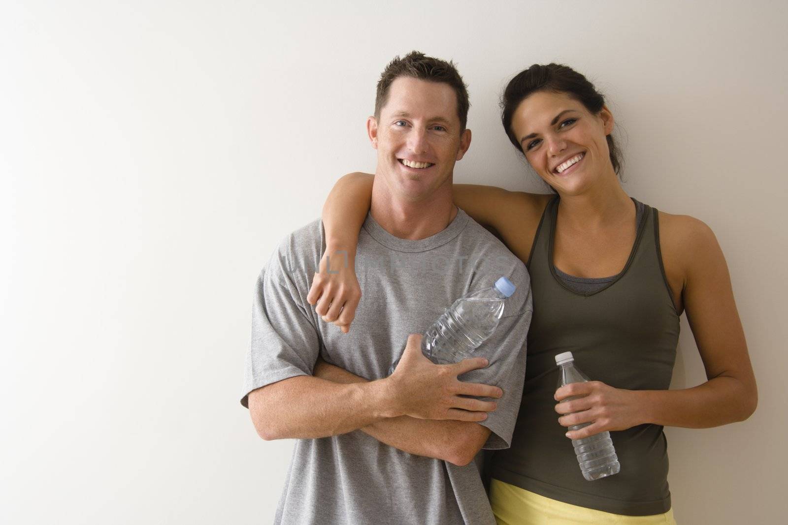 Man and woman at gym in fitness attire holding water bottles standing against wall smiling.