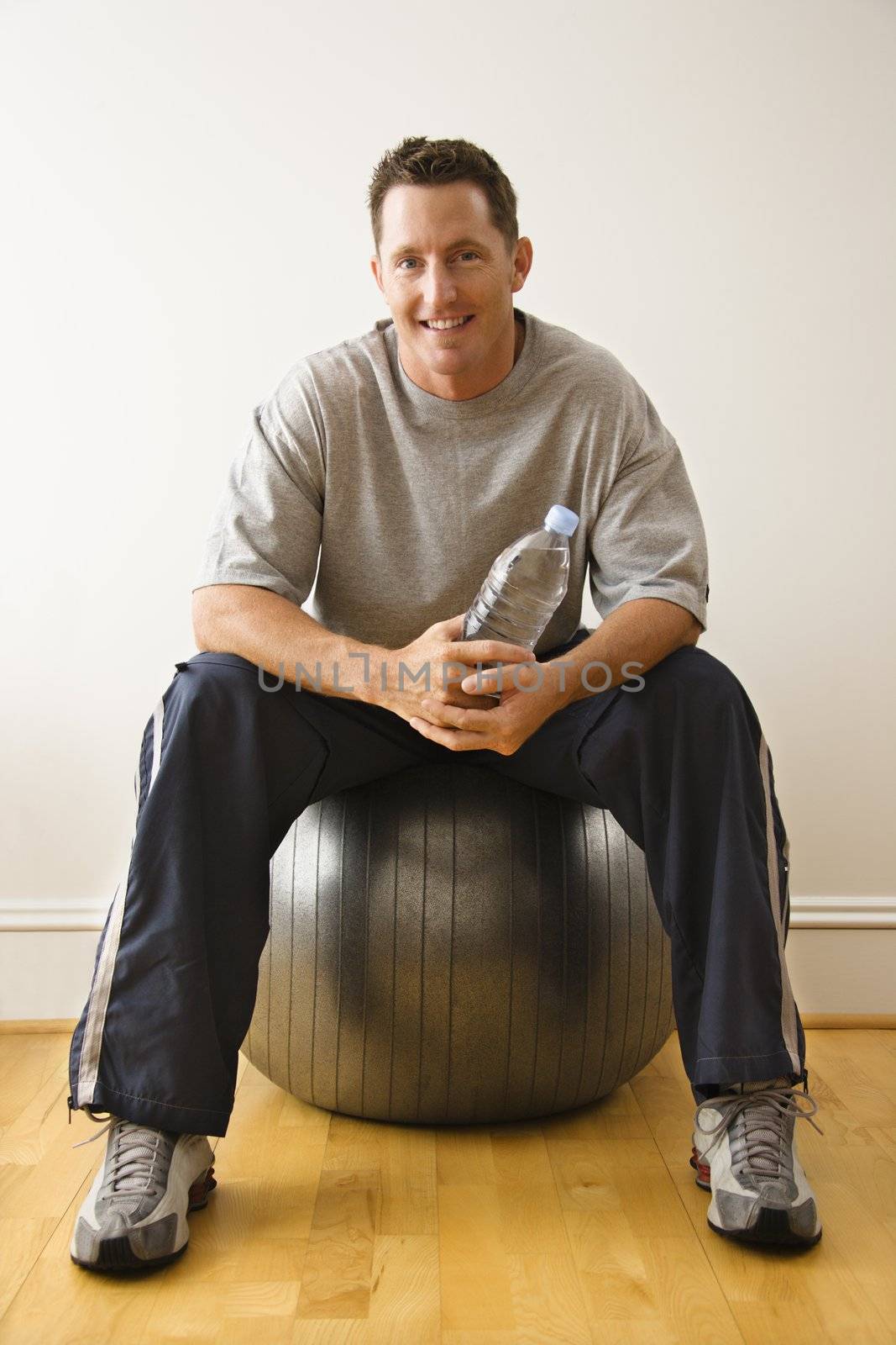 Man holding water bottle sitting on balance ball at gym smiling.