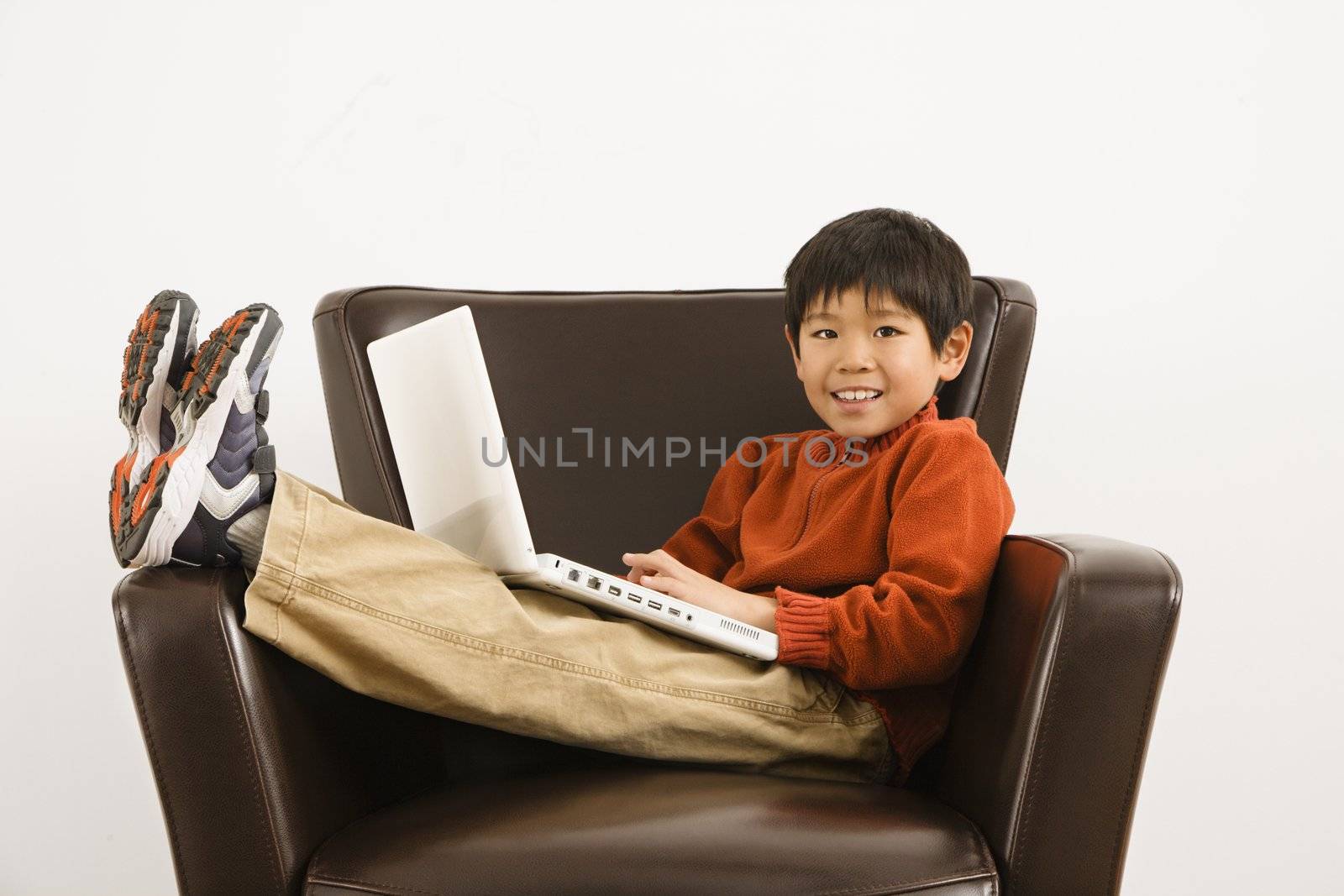 Asian boy with laptop computer sitting in chair smiling.