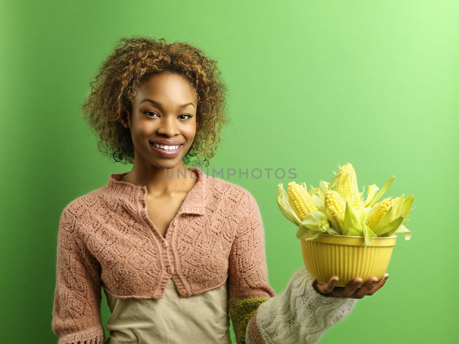 Portrait of pretty young woman standing against green background holding bowl full of ears of corn.