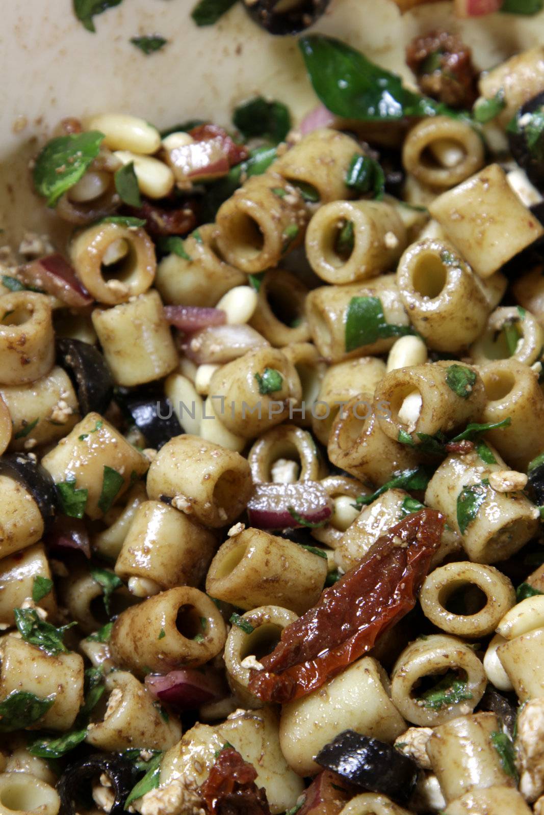 A close-up of greek pasta salad, featuring sun-dried tomatoes, black olives, red onion, pine nuts, spinach,feta cheese, balsamic vinegar and ditali pasta.

