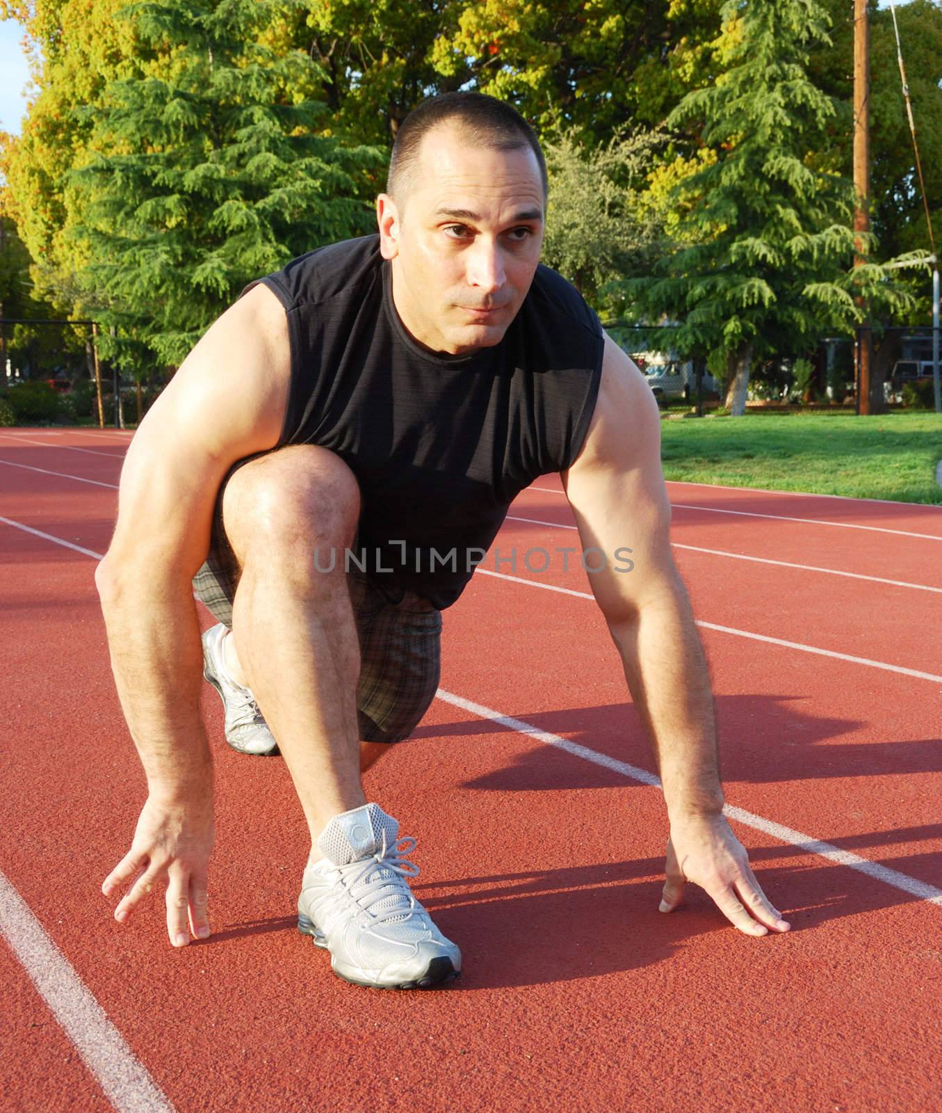 Male athlete getting ready to run on an athletic track with trees in the background on a sunny day.