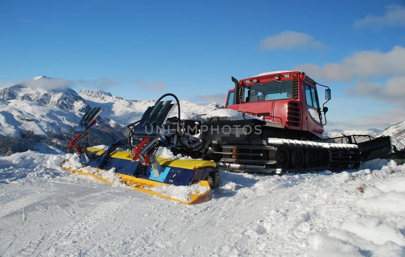 We can  see a tractor in the middle of a mountain, full of snow, in the pyrenees, in France, (el Formigal). This is the tractor who prepare the tracks for skiing.
In the background we can see mountains and the blue sky, with some grey clouds.