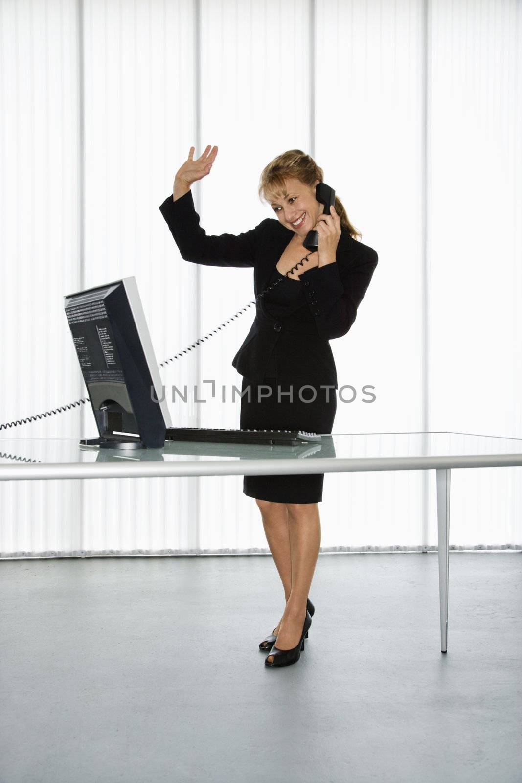 Caucasian businesswoman standing at computer desk waving on telephone.