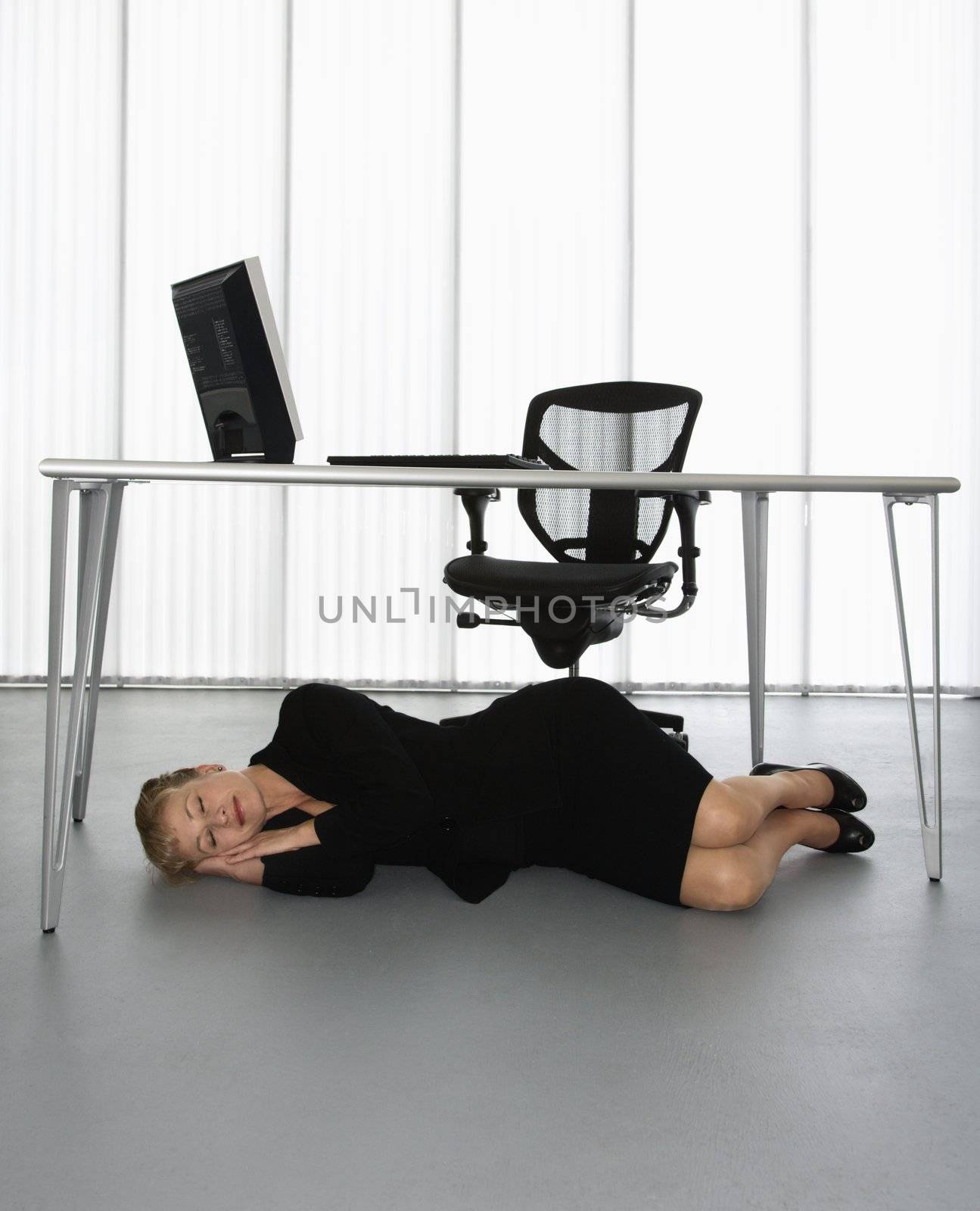 Caucasian businesswoman sleeping on floor under computer desk.