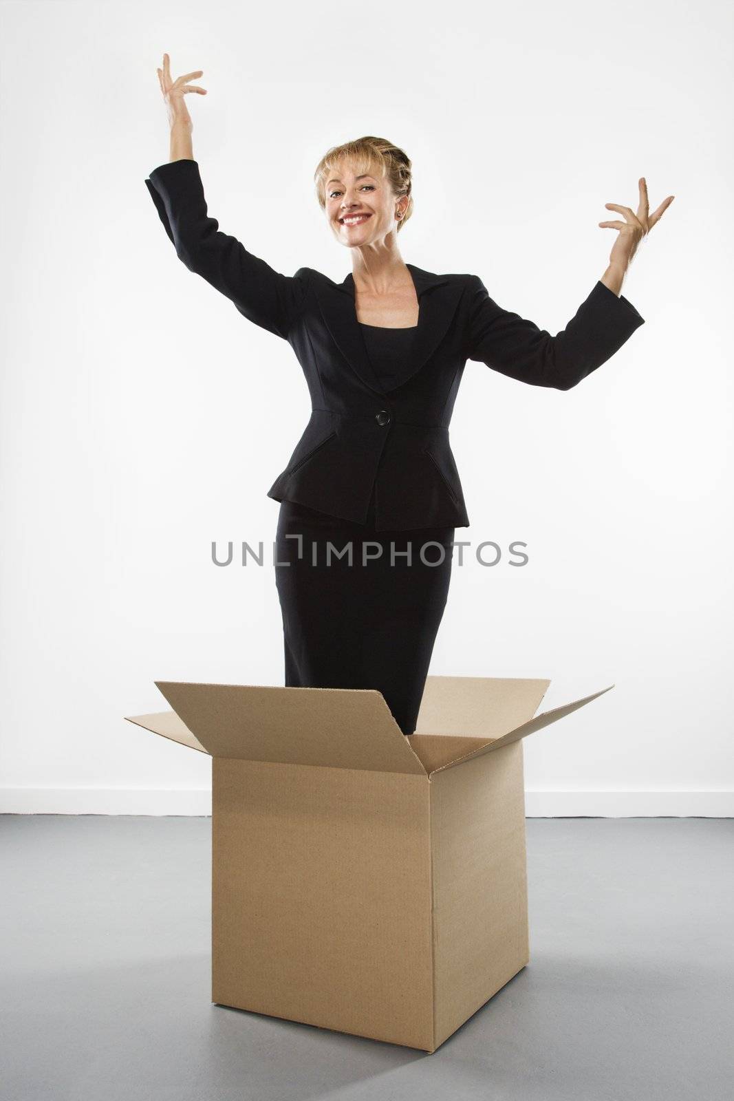 Caucasian businesswoman standing in cardboard box gesturing above head and smiling at viewer.