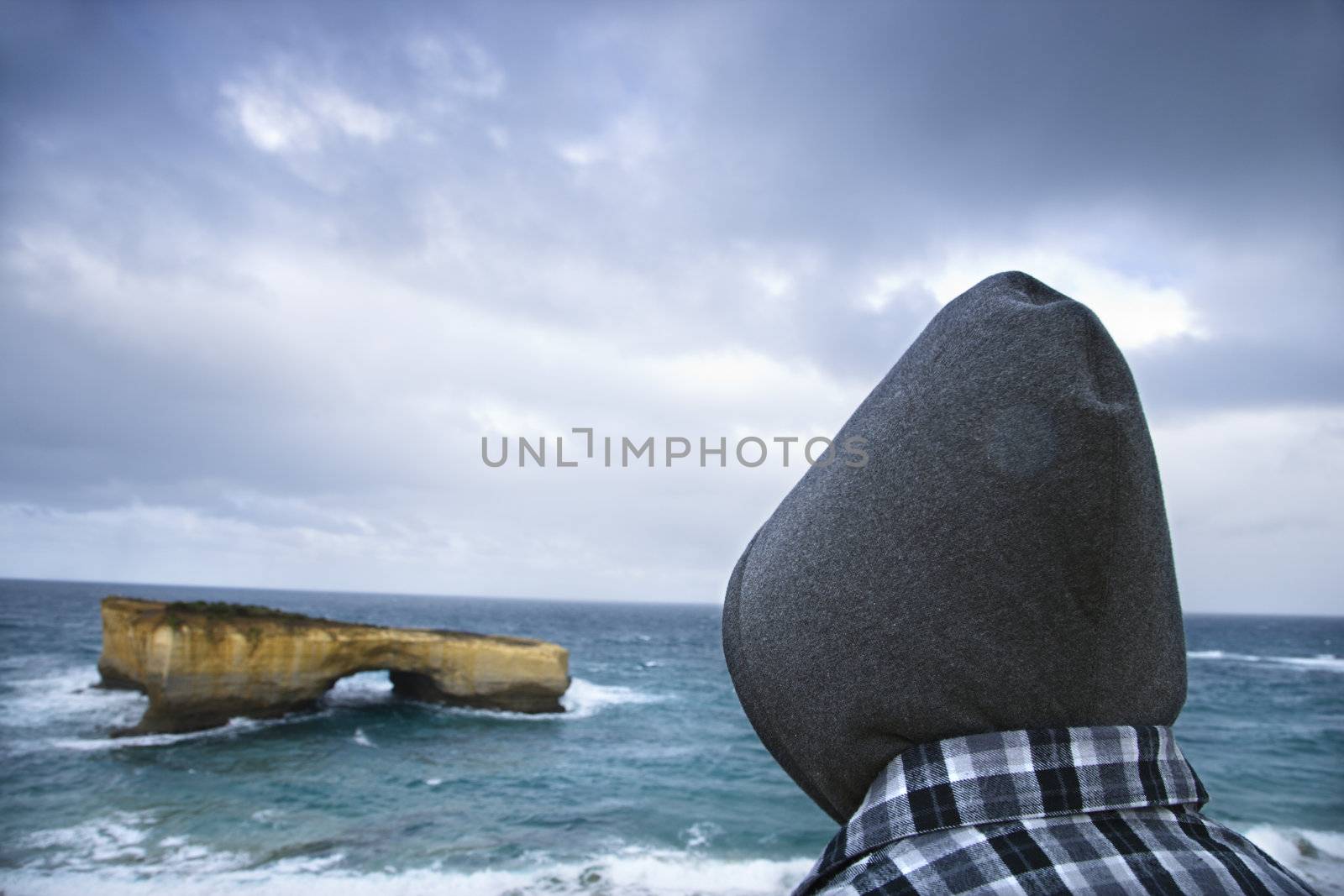 Back view of person wearing hooded clothing facing rock formation in arch shape as seen from Great Ocean Road in Australia.