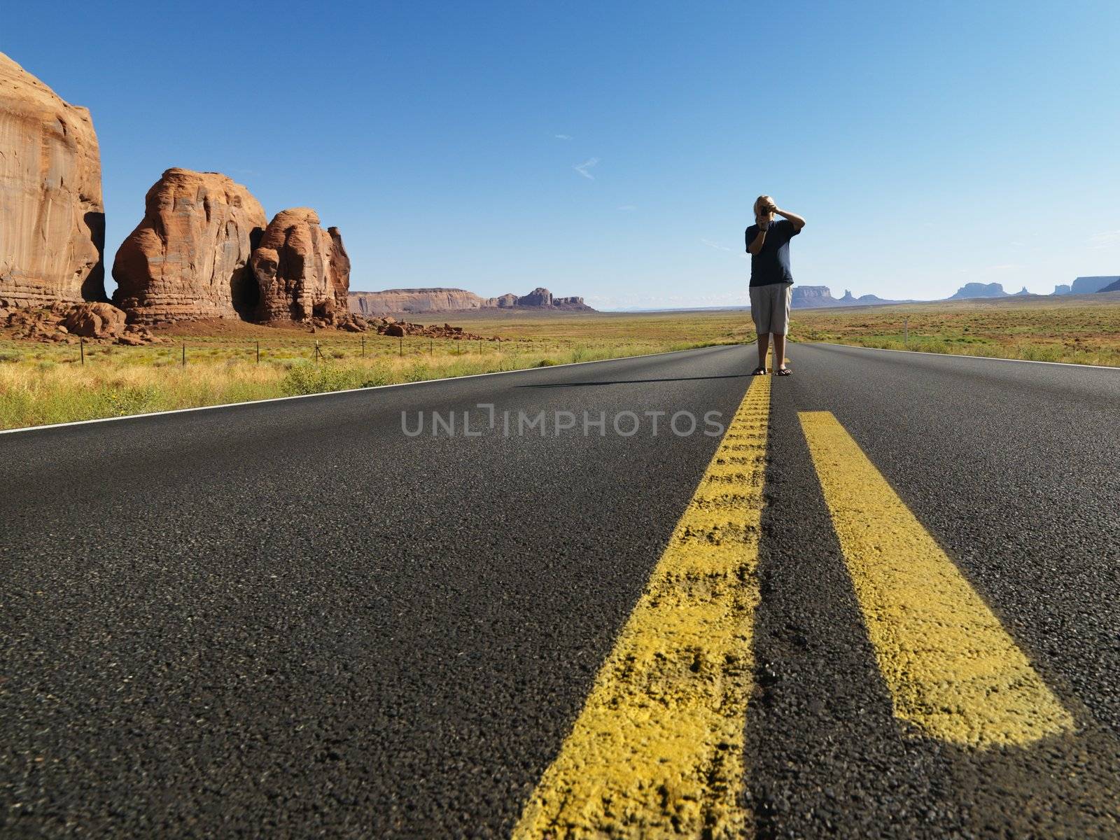 Caucasian teenage boy standing in open road in desert with landforms taking photograph.