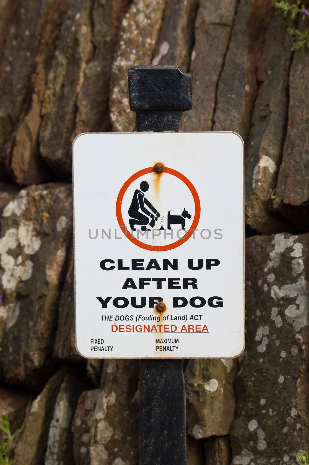 an old, worn, rust stained dog fouling sign on a black post in front of a stone wall, with fixed and maximum penalty removed.