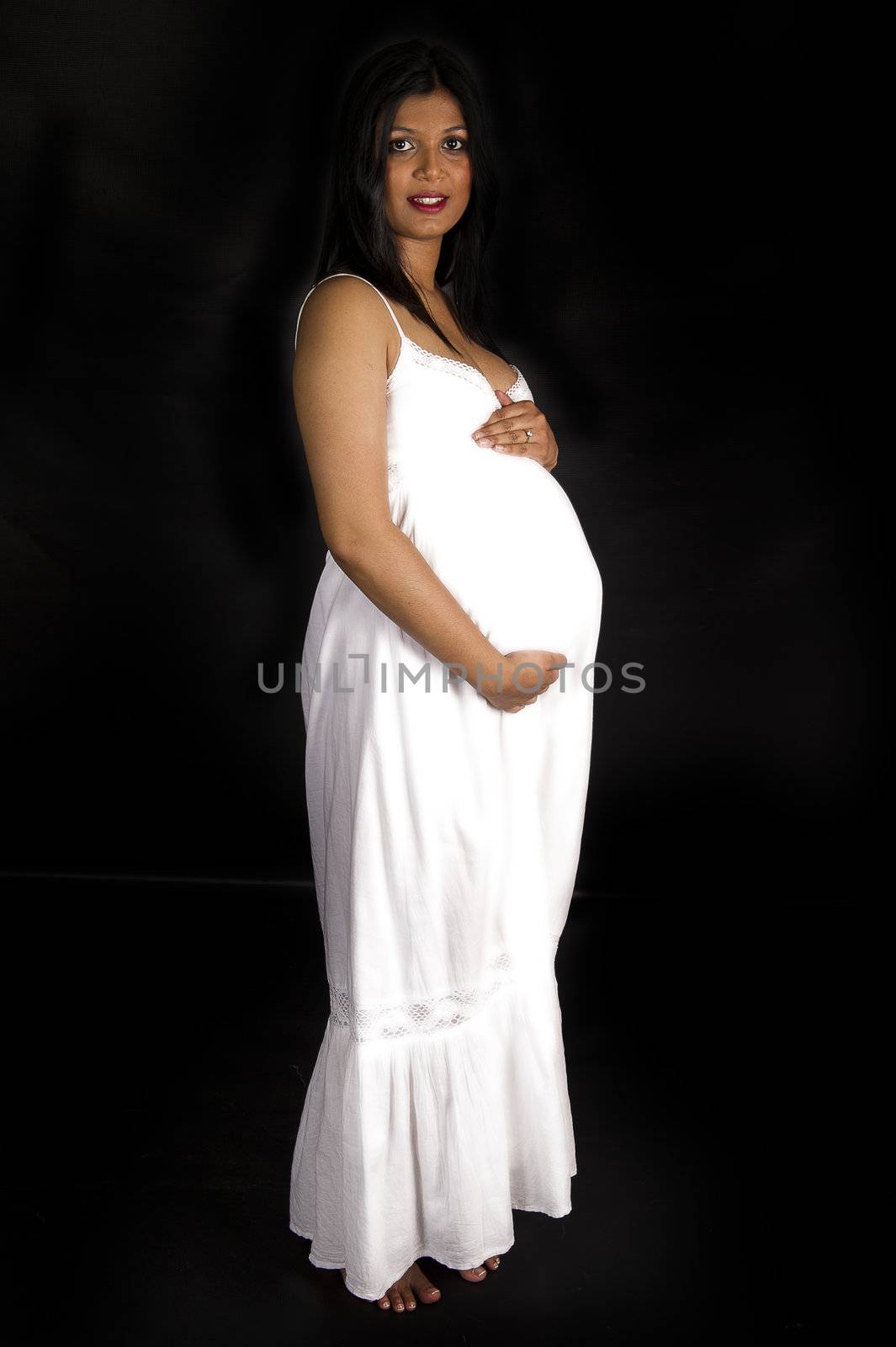 A beautiful pregnant Indian woman in white dress and angel wings smiling on black backdrop