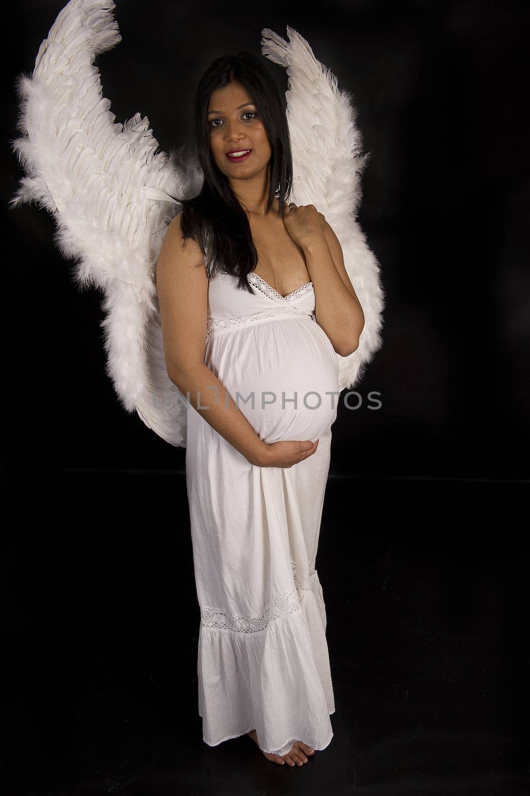 A beautiful pregnant Indian woman in white dress and angel wings smiling on black backdrop