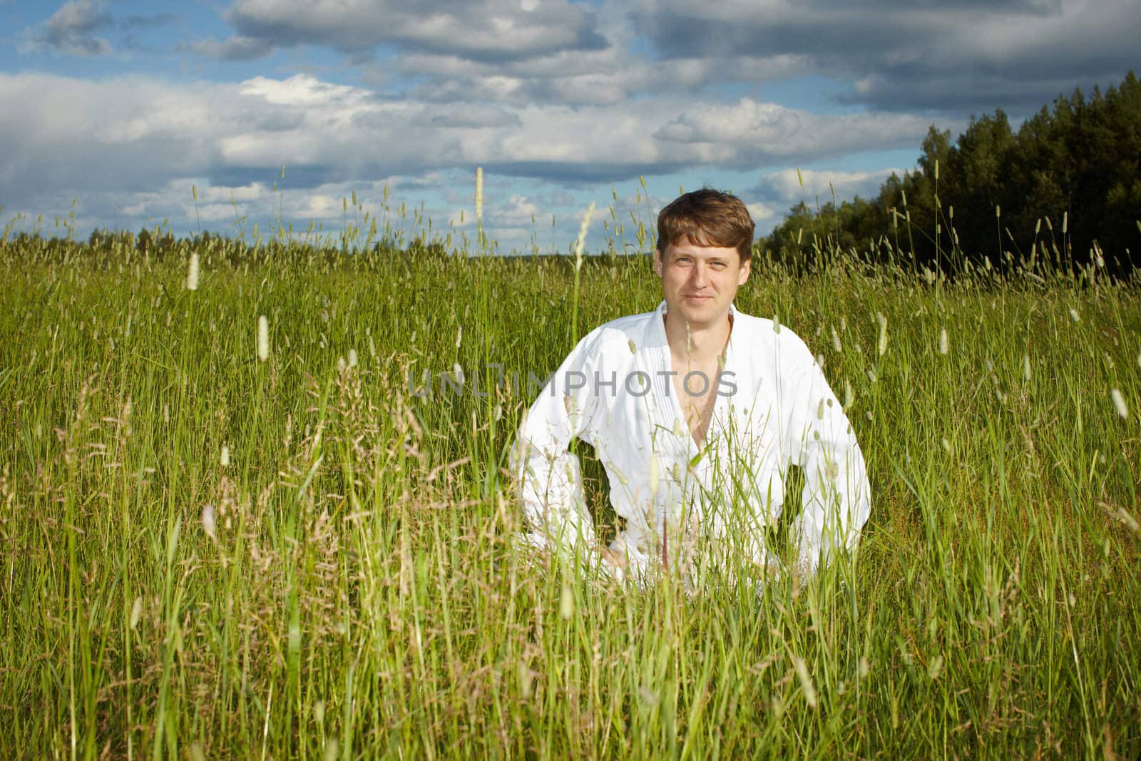 Young man in white kimono sits in grass by pzaxe