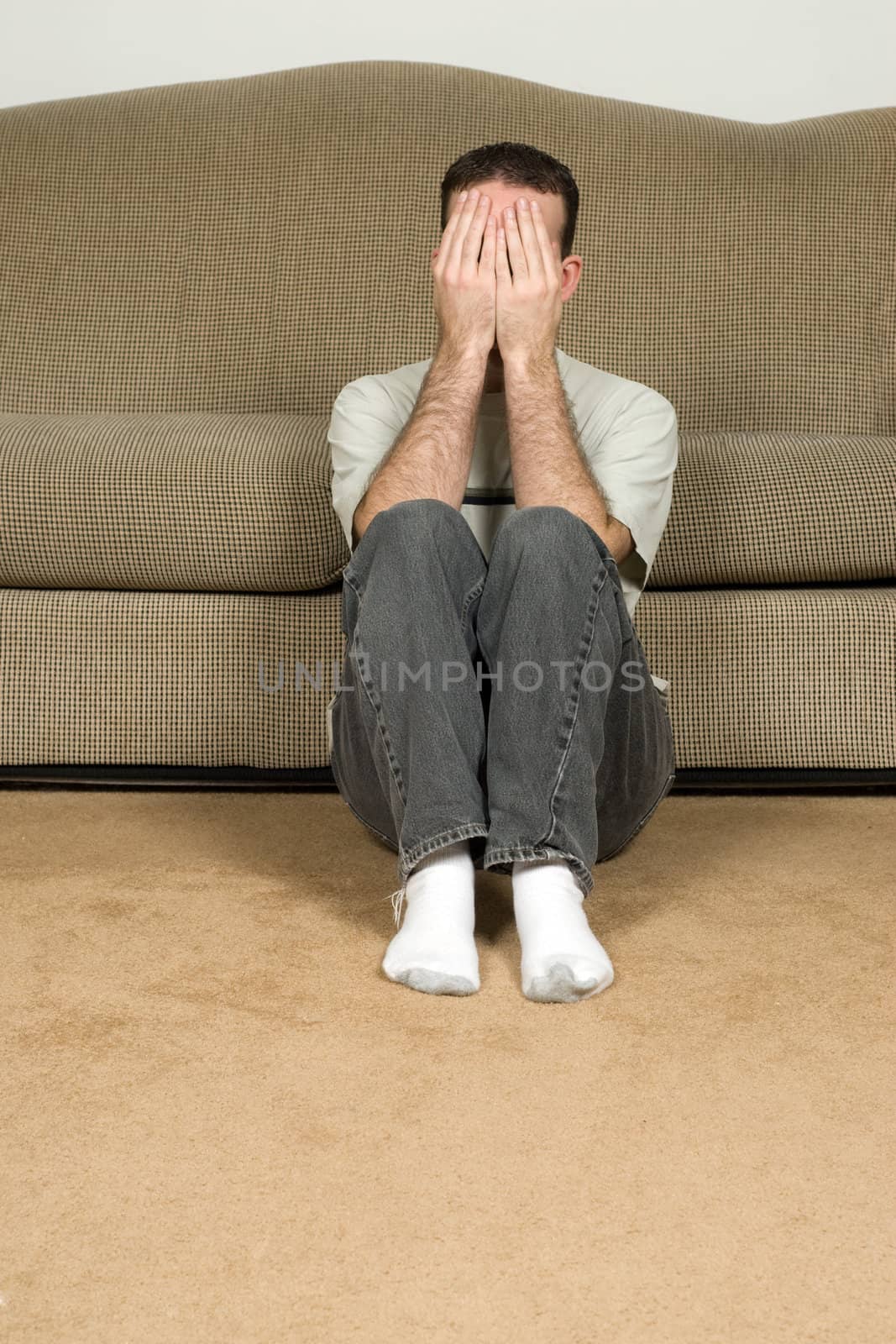 A depressed man covering his face and sitting on the floor near the sofa