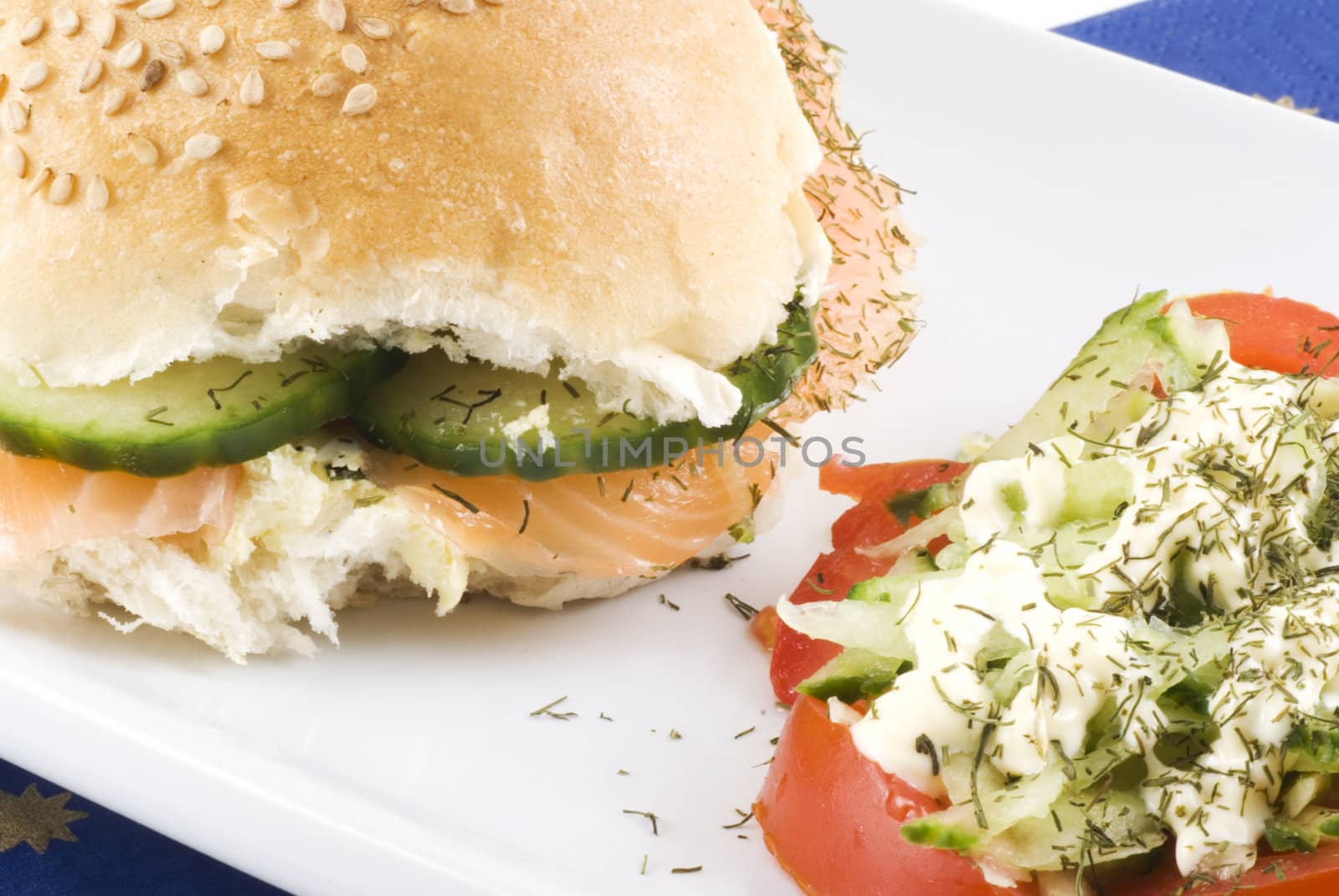 Close up of a sesame bun with salmon and cucumber with a tomato salad on the side.