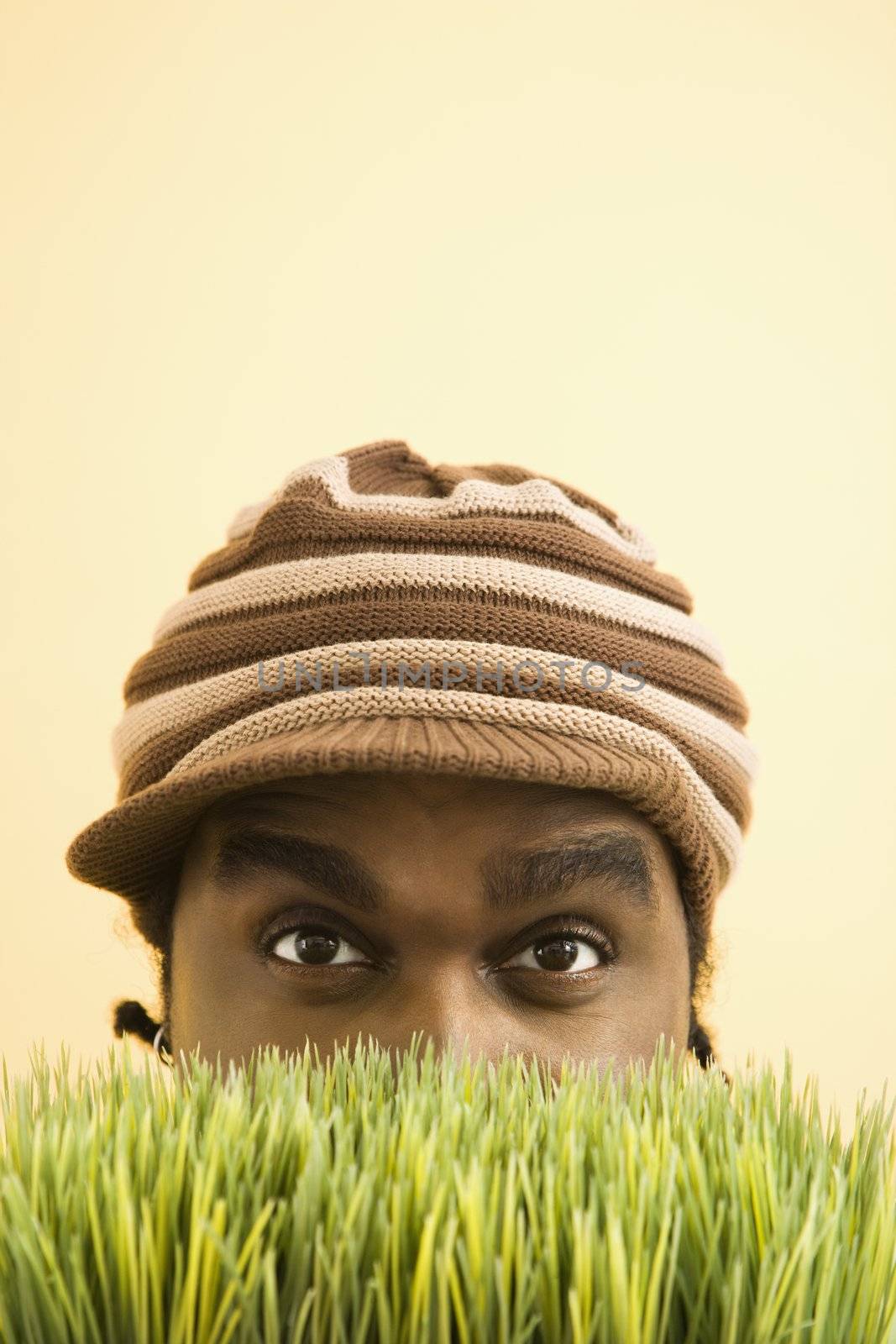 African-American mid-adult man wearing knit hat with brim peeking at viewer from behind grass.