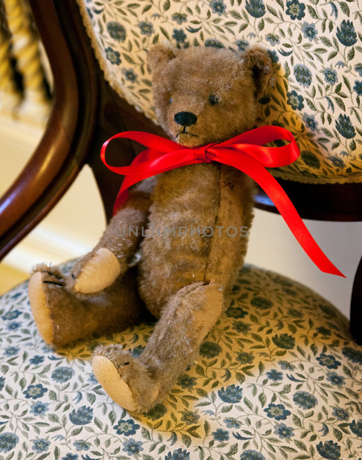 Old teddy bear on ornate seat with red ribbon around neck and missing one arm