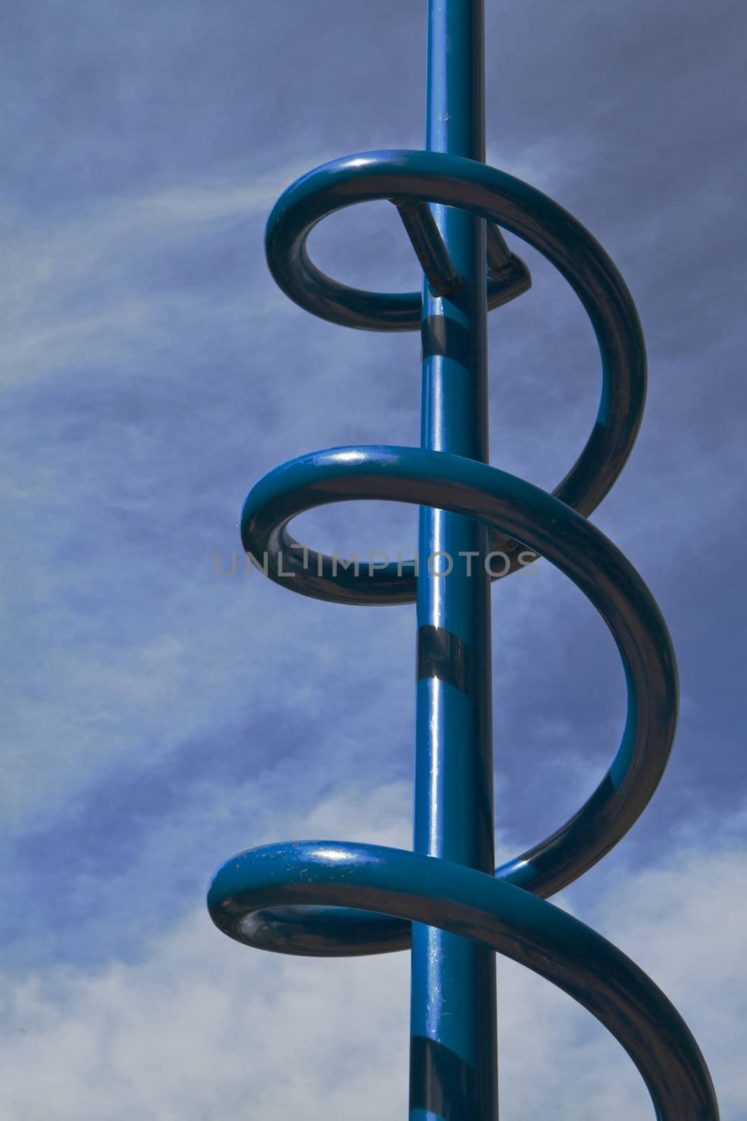 Sun lit blue metal climbing spiral feature of a childrens playground with cloudscape background