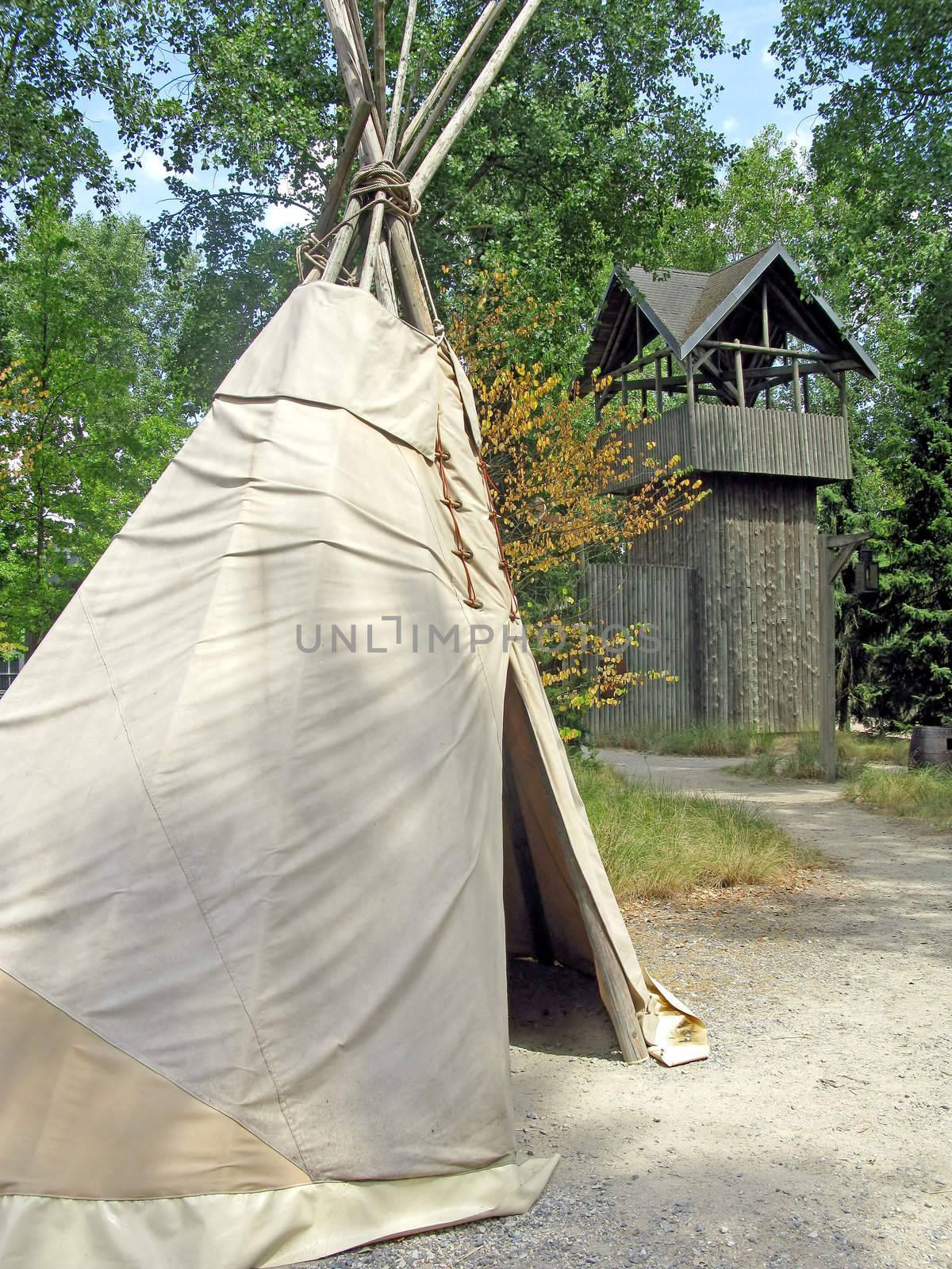 Teepee in a forest with a wooden fort behind.