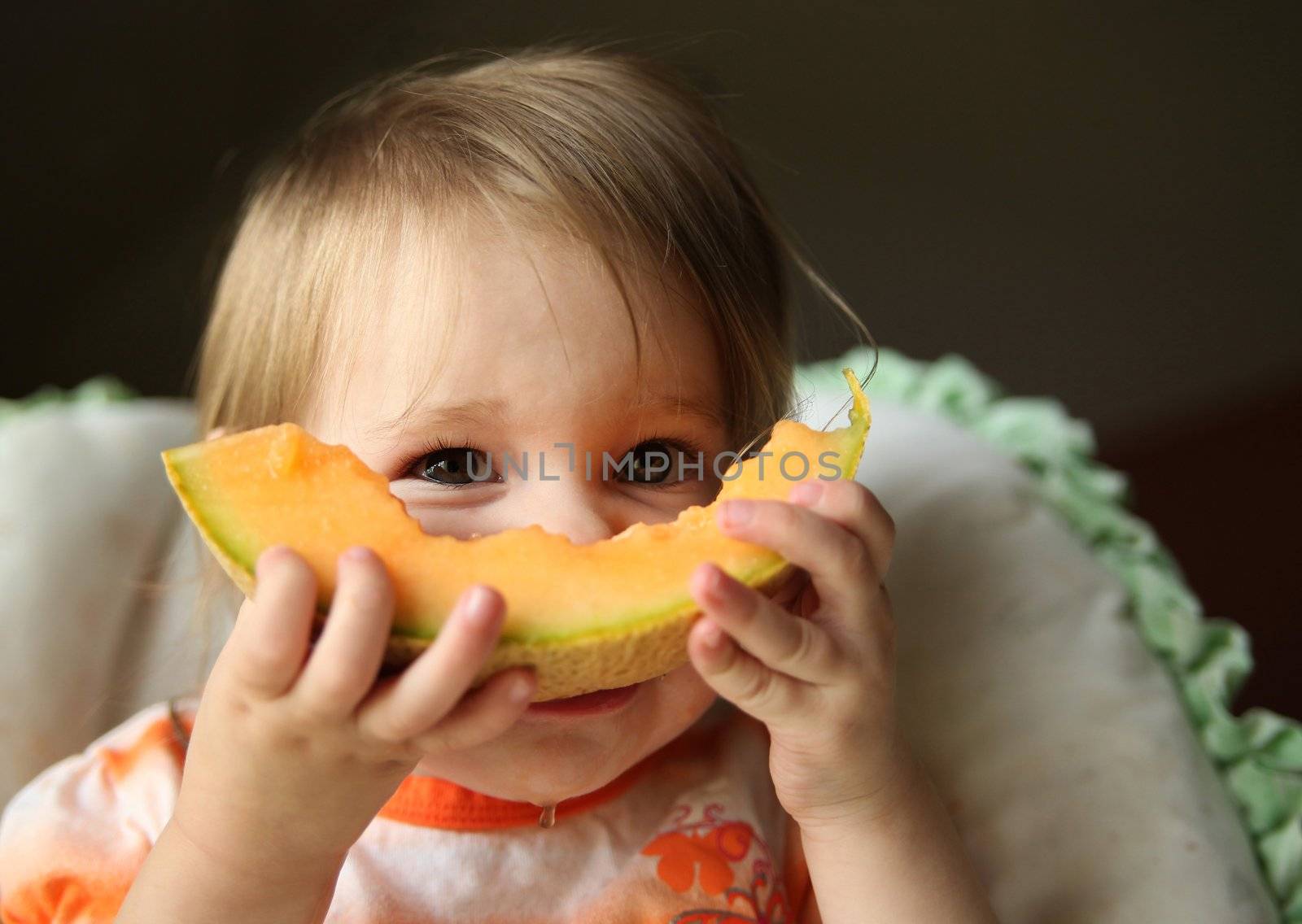 Young baby girl sitting in a high chair eating a wedge of cantaloupe with juice dripping off her chin.