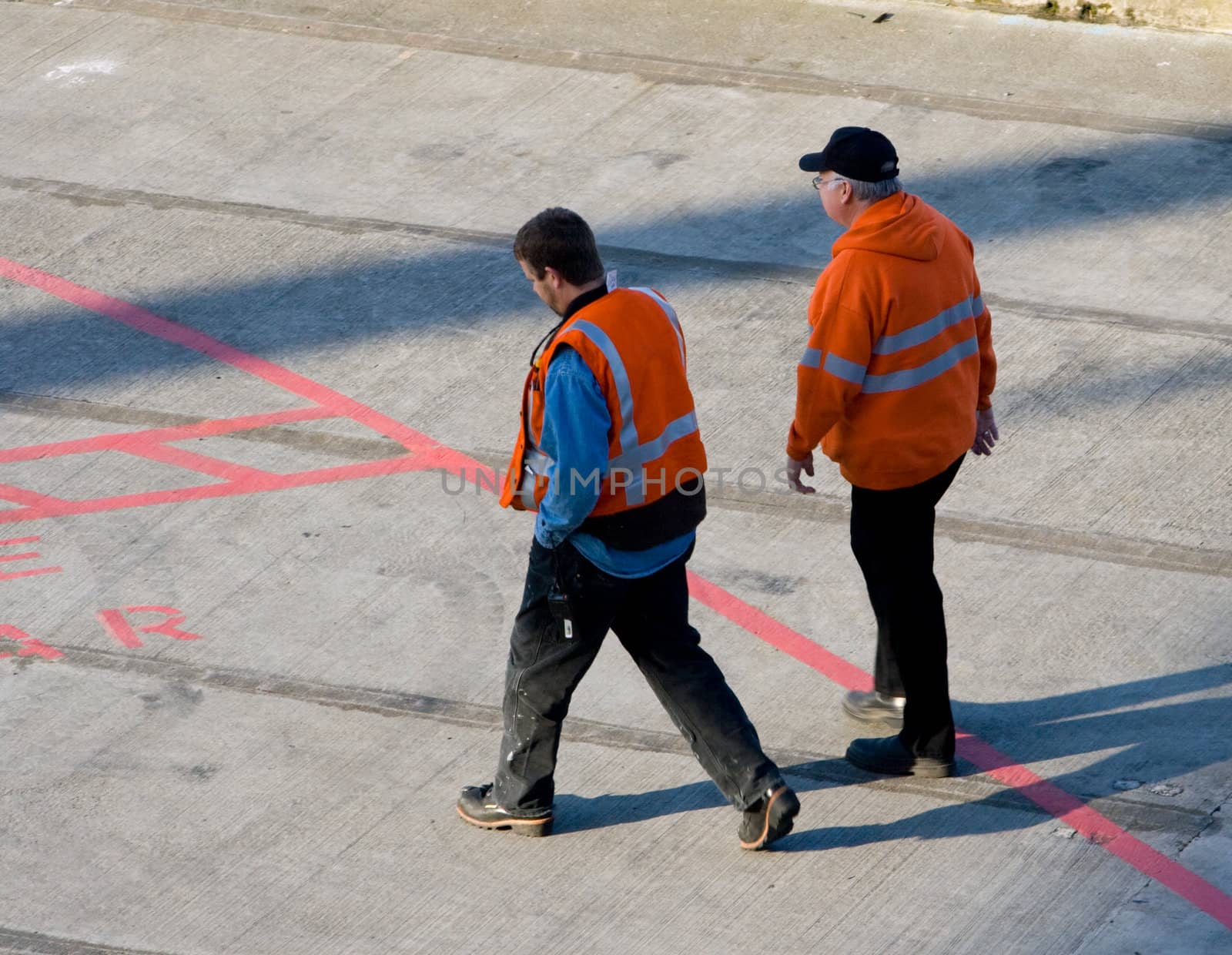 Pier Workers with Orange Vests v1 are two men that are walking along the dock areas which border Puget Sound and the port of Seattle Washington. One man is wearning a hate and both have reflective strips and are wearing black pants.
